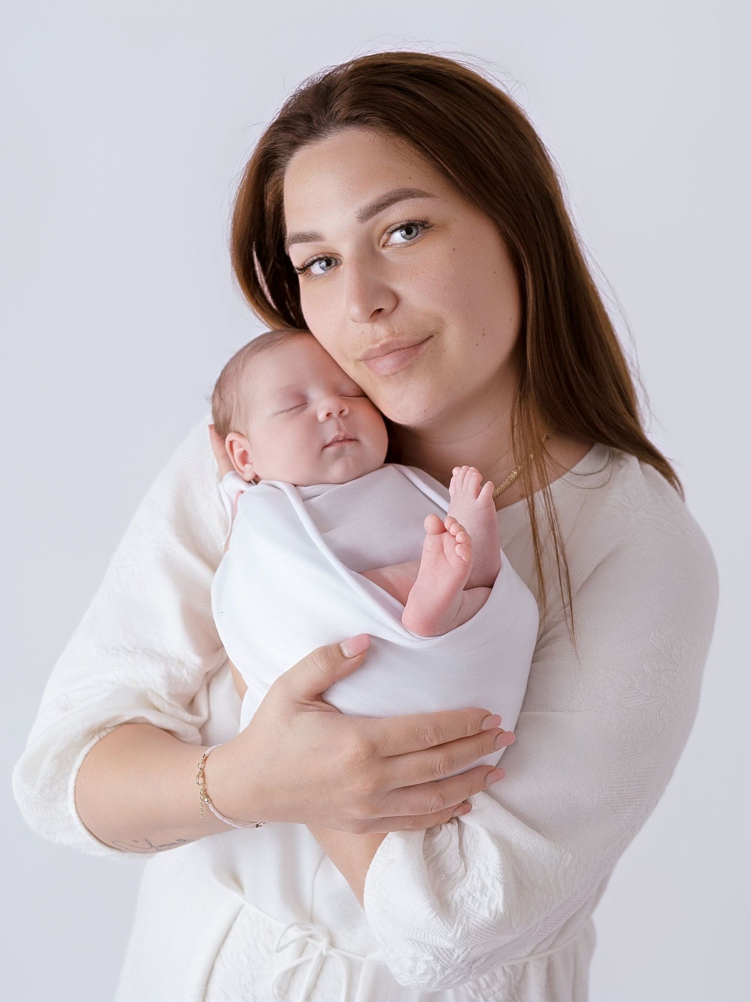 Young mother in white dress tenderly holds sleeping newborn baby wrapped in white swaddle against light background.