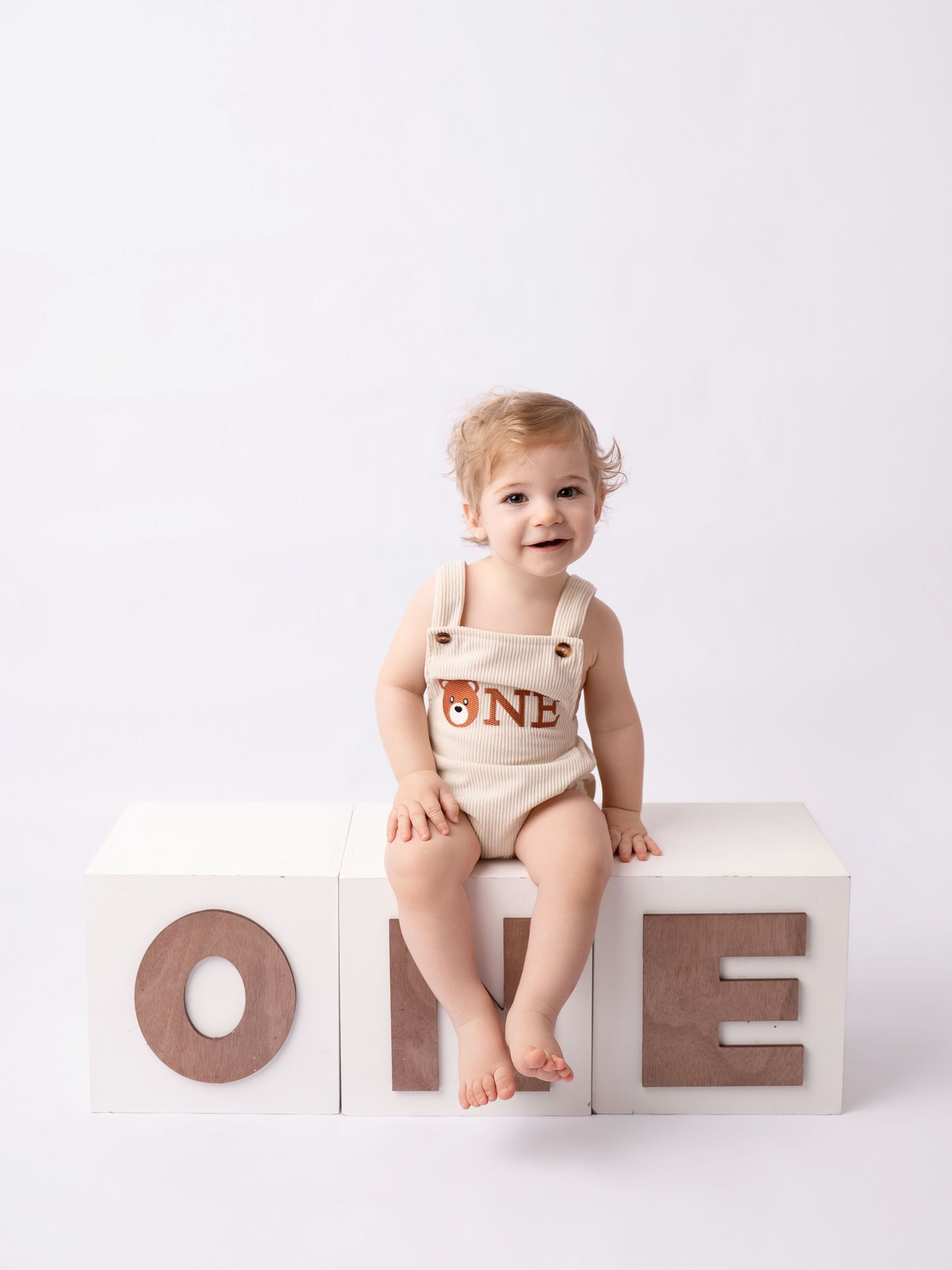 Cute baby in bear overalls sitting on ONE letter blocks for first birthday photos.