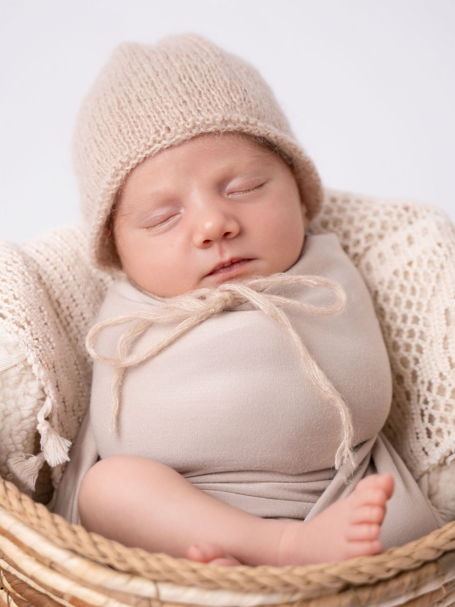 Sleeping newborn baby wrapped in beige swaddle with knit bonnet in a wicker basket.
