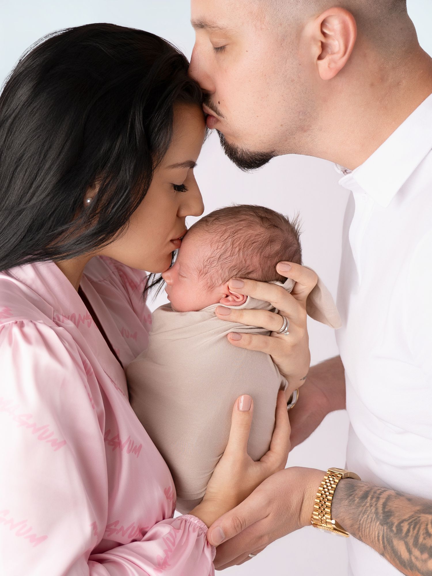 A couple tenderly kisses their swaddled newborn baby, the mother in pink and father in white, against a soft blue background.