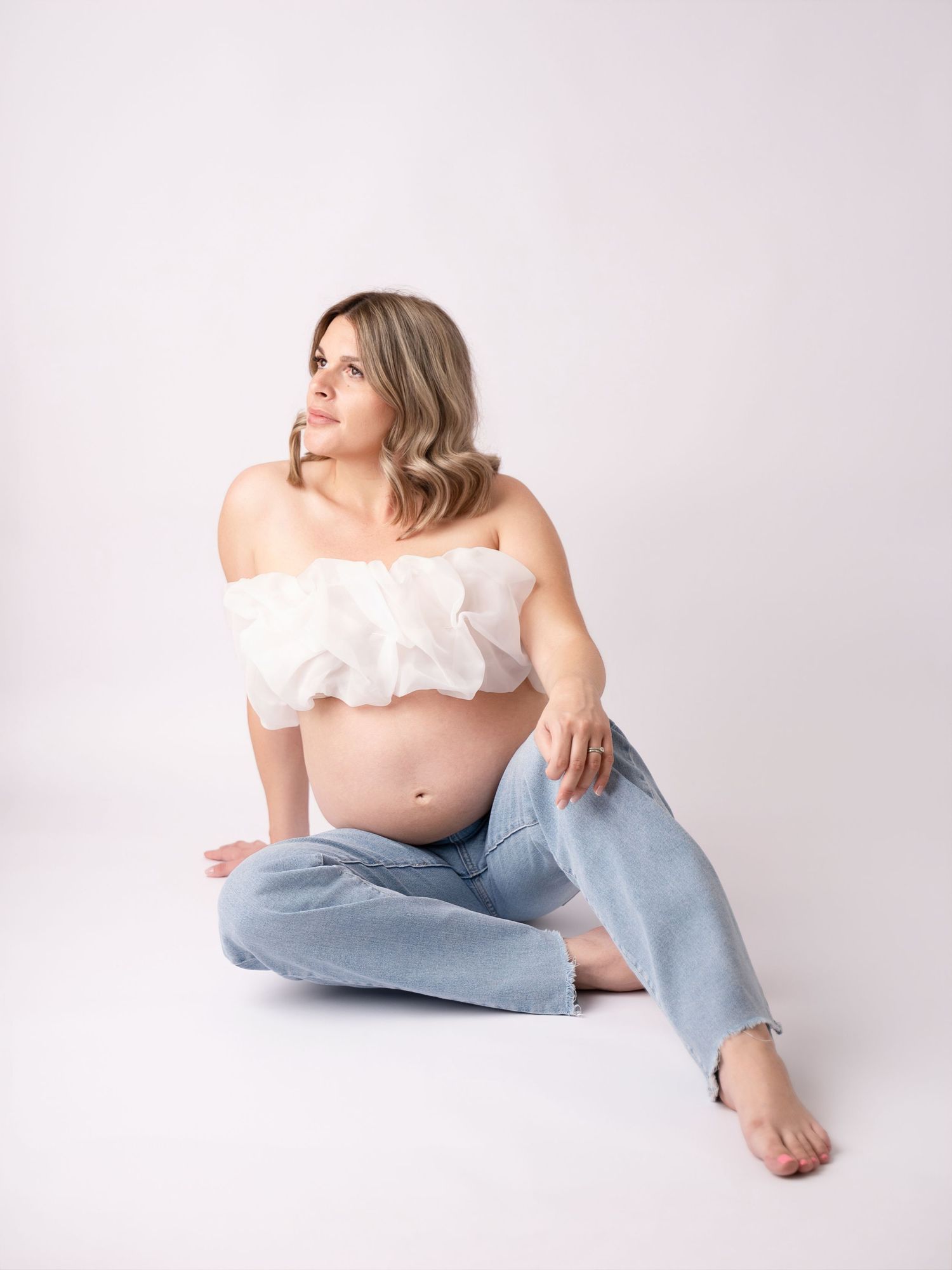Pregnant woman in white ruffled tube top and light blue jeans sitting on floor, looking sideways in studio.