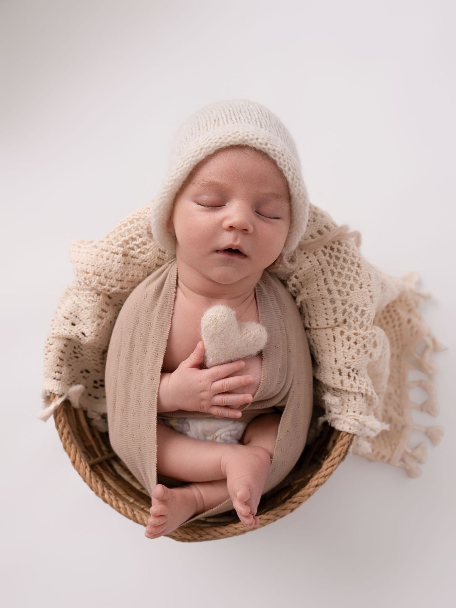 Sleeping newborn baby in a wicker basket wearing a cream knit bonnet, holding a heart toy, wrapped in soft beige fabric.