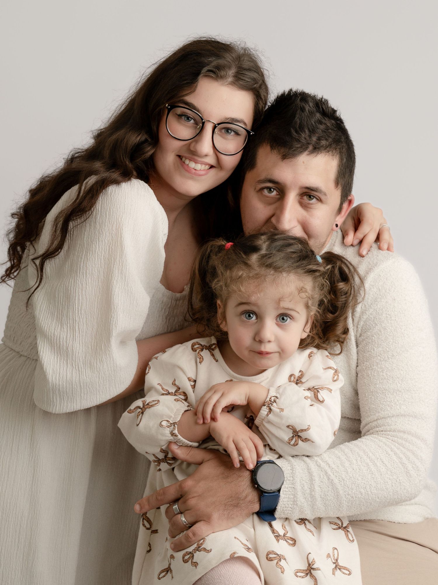 Happy family portrait with mother, father, and young daughter in coordinating cream outfits on white background.