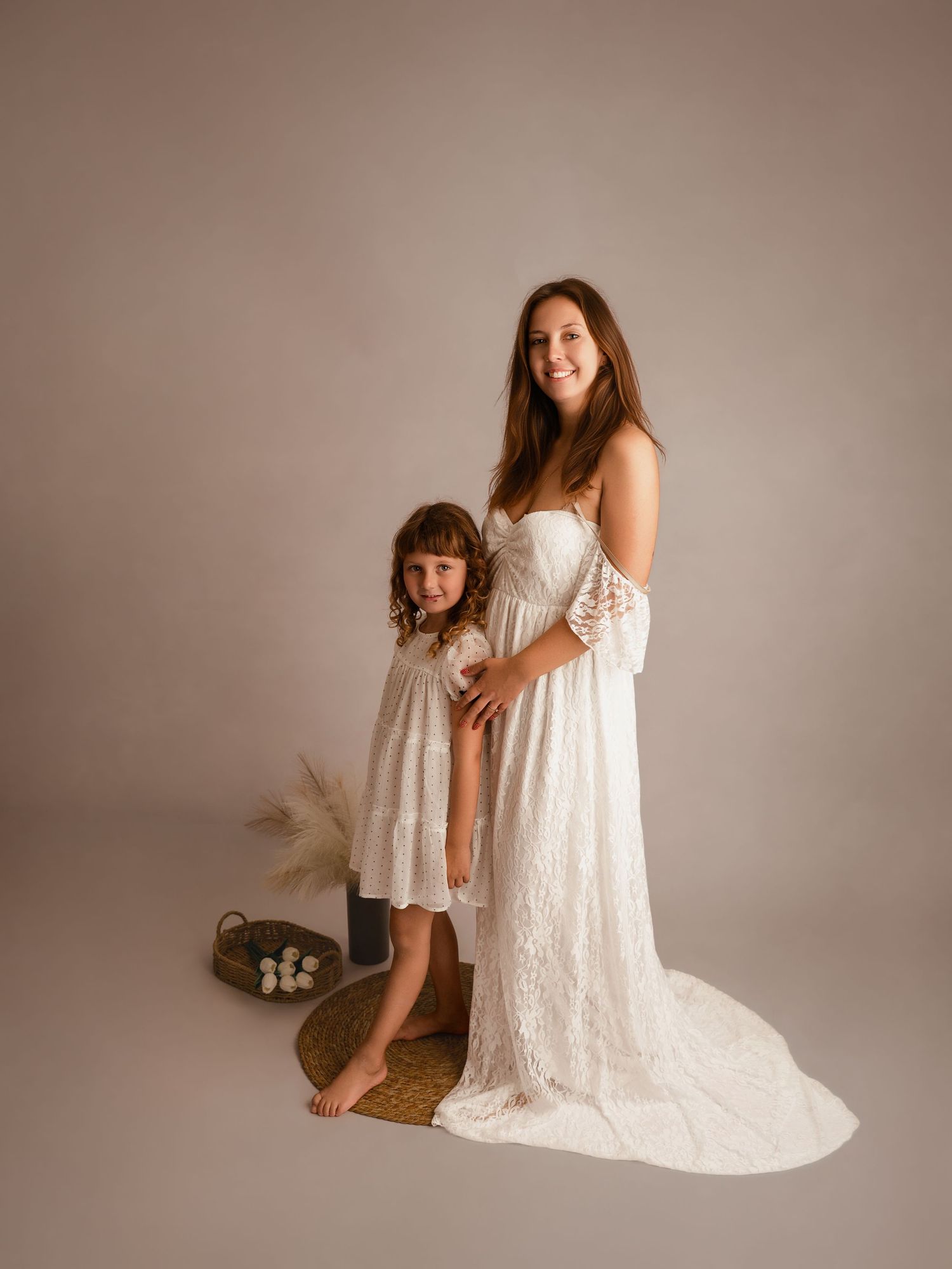 Pregnant woman in white lace gown poses with young girl in matching white dress in studio photo.
