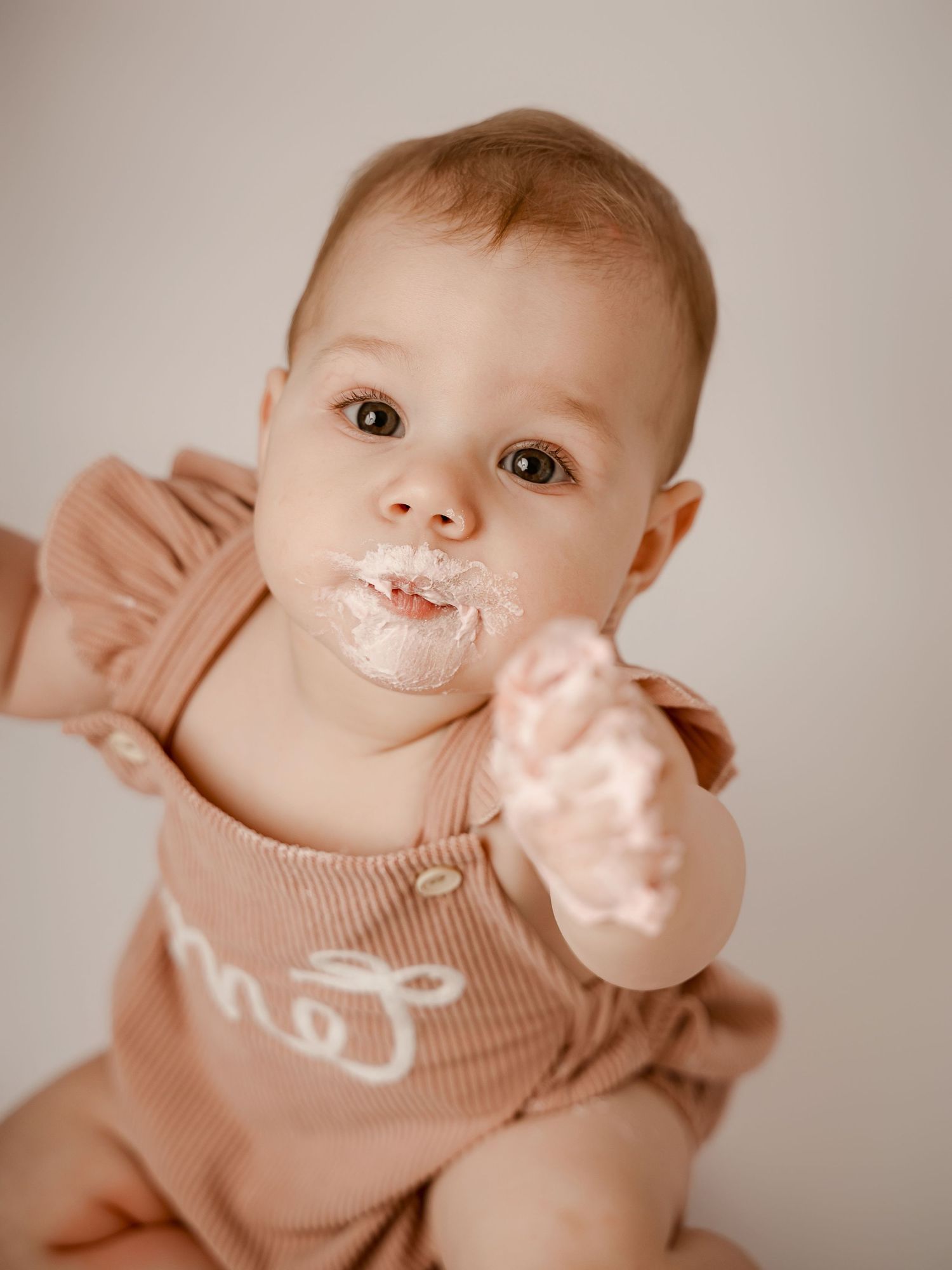 Cute baby with frosting on face wearing pink ribbed romper, reaching out during first birthday cake smash photo session.