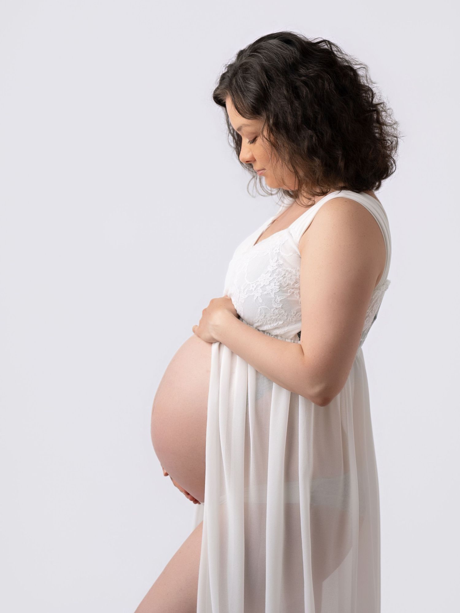 Pregnant woman in white lace dress cradling baby bump in studio maternity photo session.