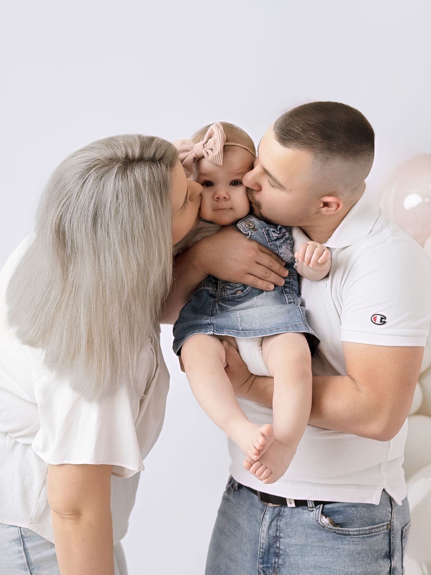 A couple in white tops kisses their baby girl in a denim outfit during a studio family photo session.