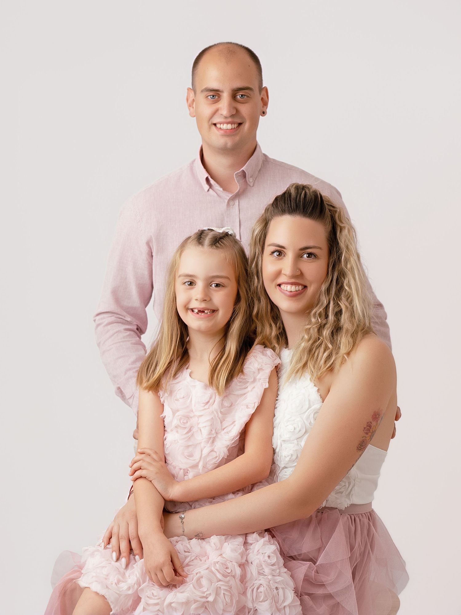 A smiling family of three in coordinating pink outfits poses together for a professional portrait against a light background.