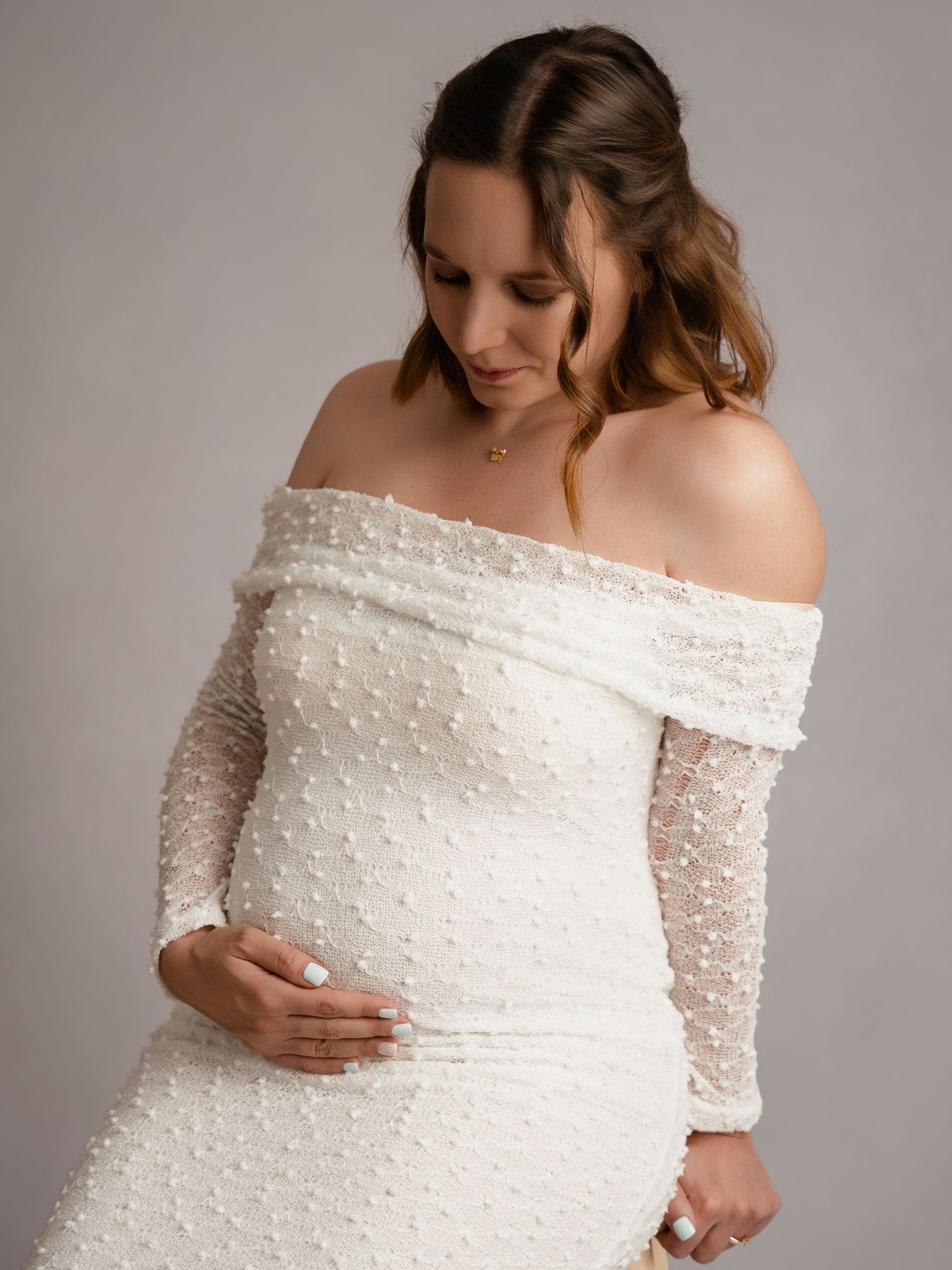 A pregnant woman in an elegant off-shoulder white lace maternity gown gently cradles her baby bump in a studio portrait.