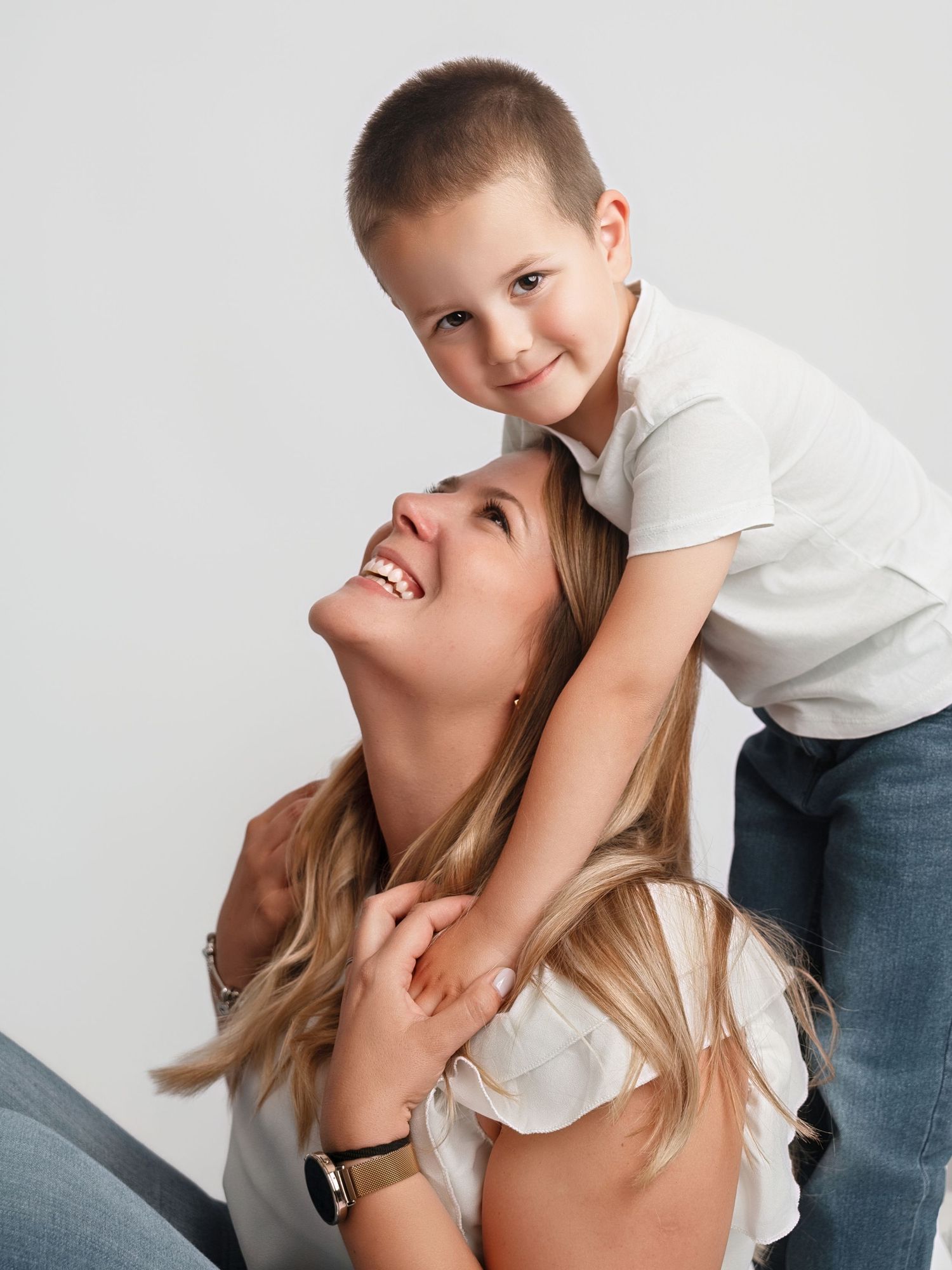 Joyful boy climbs on his laughing mothers back during a white background studio photo session.