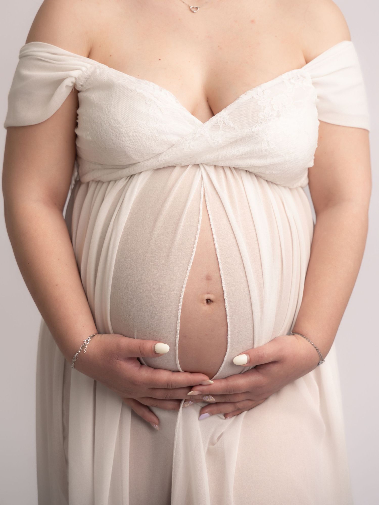 Close-up of pregnant woman in sheer white off-shoulder gown gently holding bare baby bump with both hands.