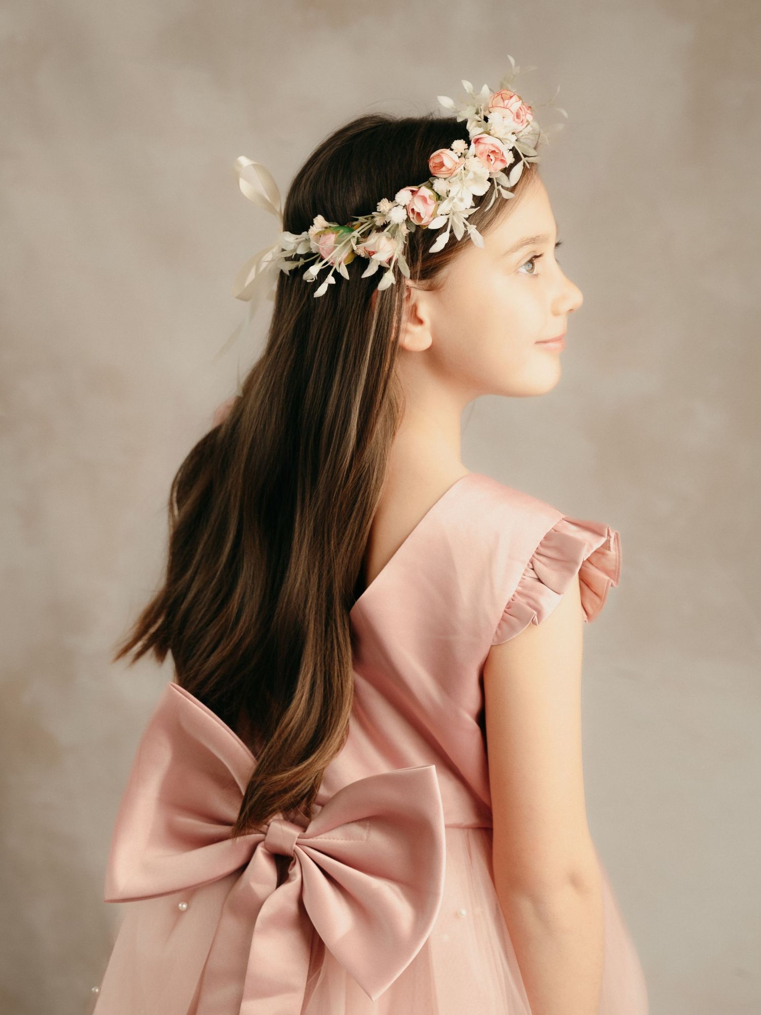 Side profile of girl in pink satin dress with ruffle sleeves and floral crown in studio setting.