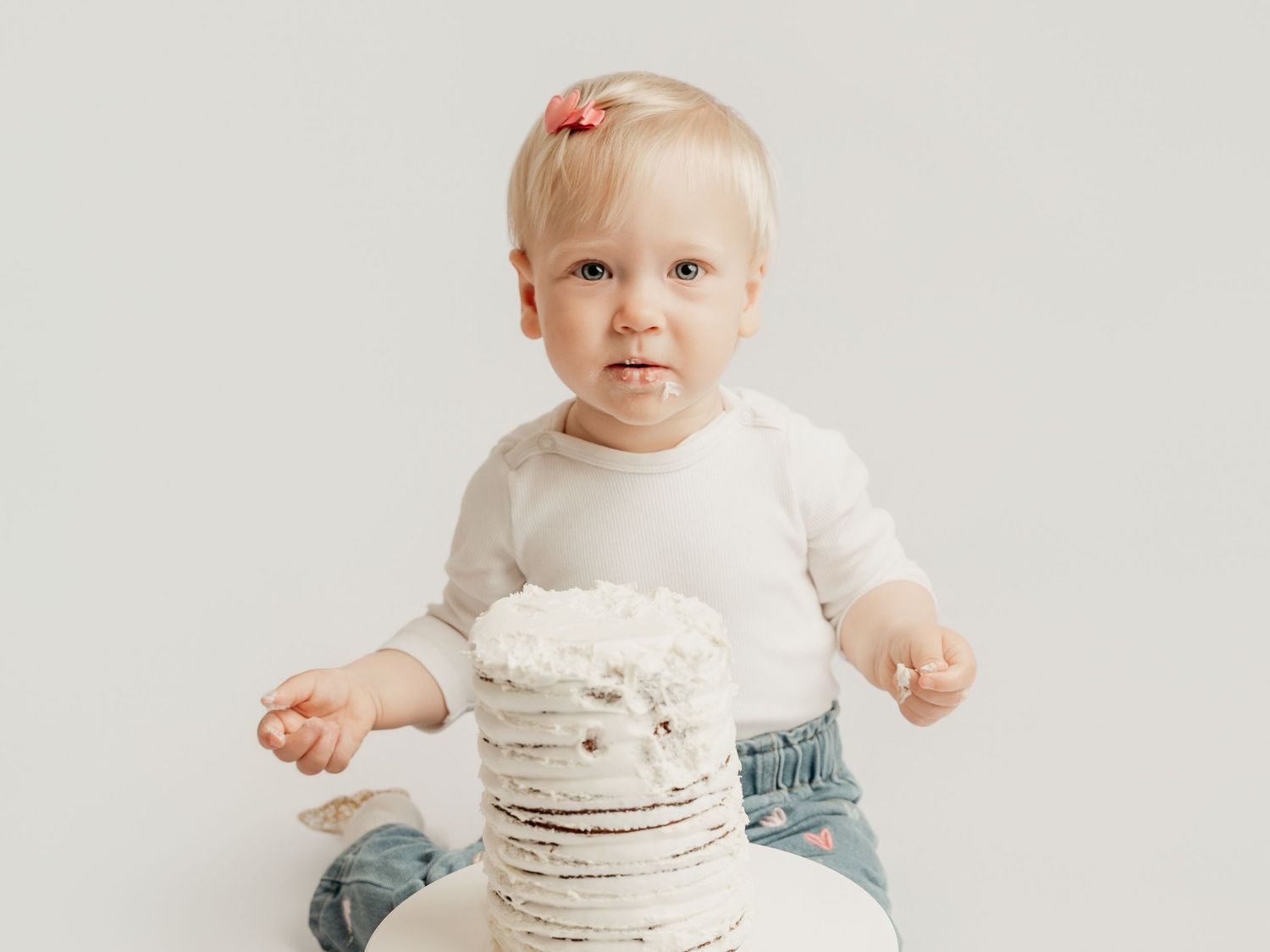Adorable baby girl in white onesie and jeans sits in front of a rustic white smash cake.