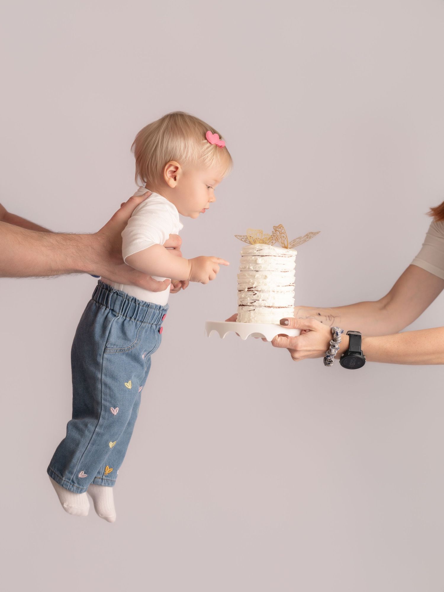 Baby girl suspended mid-air by parents reaching toward a white smash cake in birthday photoshoot.