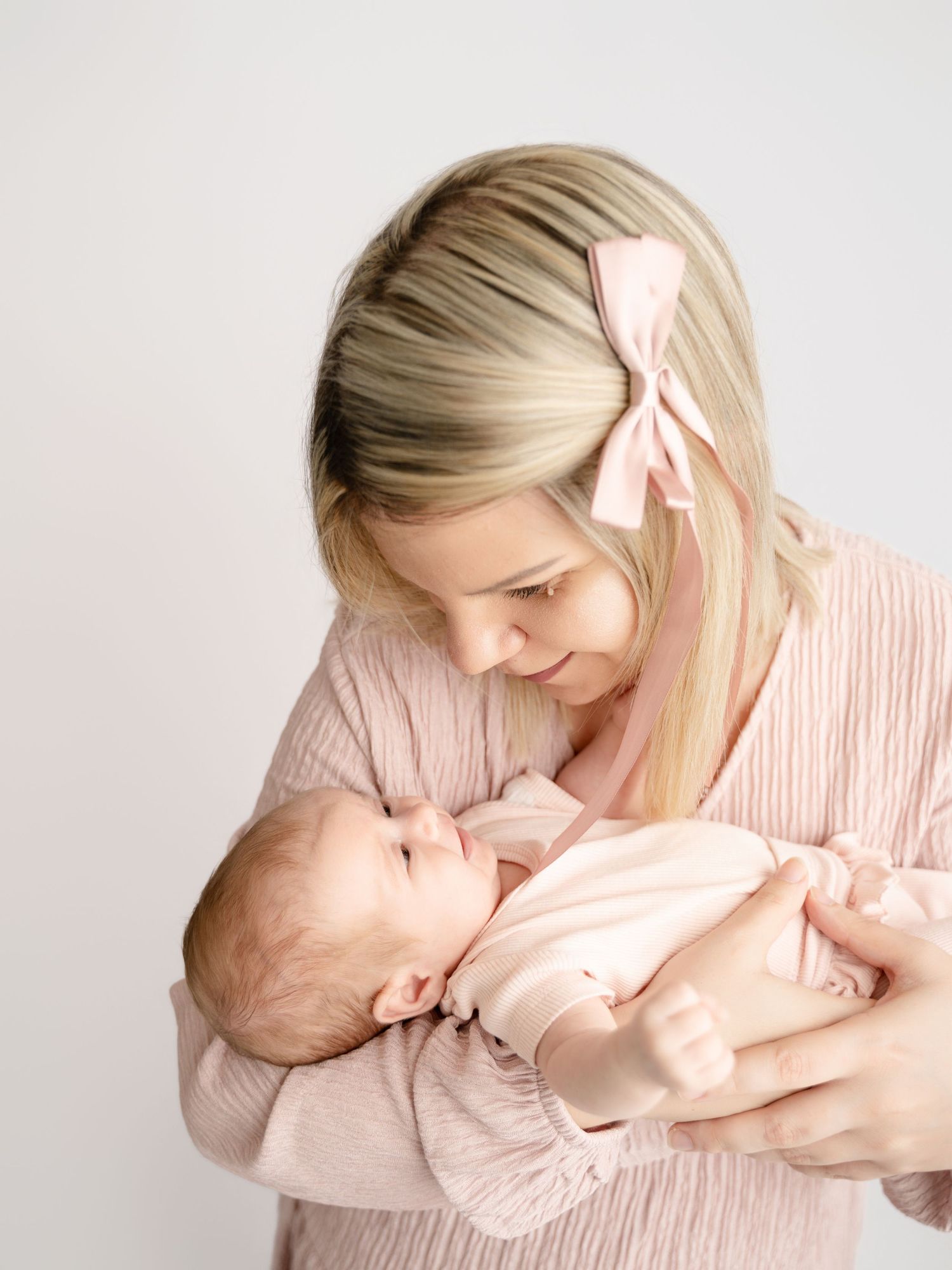 Blonde mother in pink outfit with bow headband tenderly holding newborn baby in white studio setting.