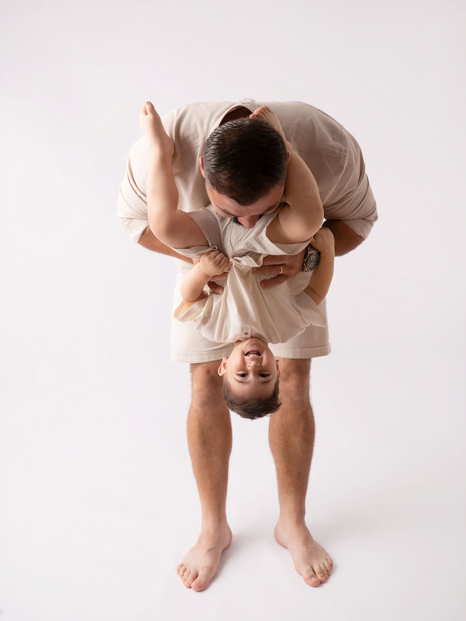 Father playfully holds toddler upside down while kissing them during a studio photo session.