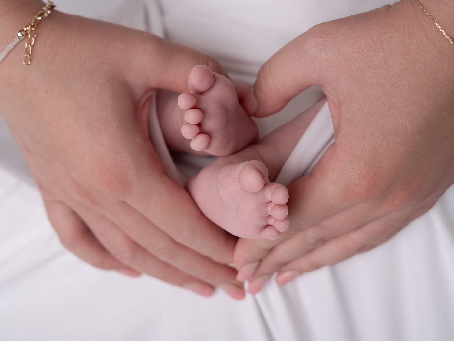 Adult hands forming a heart shape cradling tiny newborn baby feet on a white background.