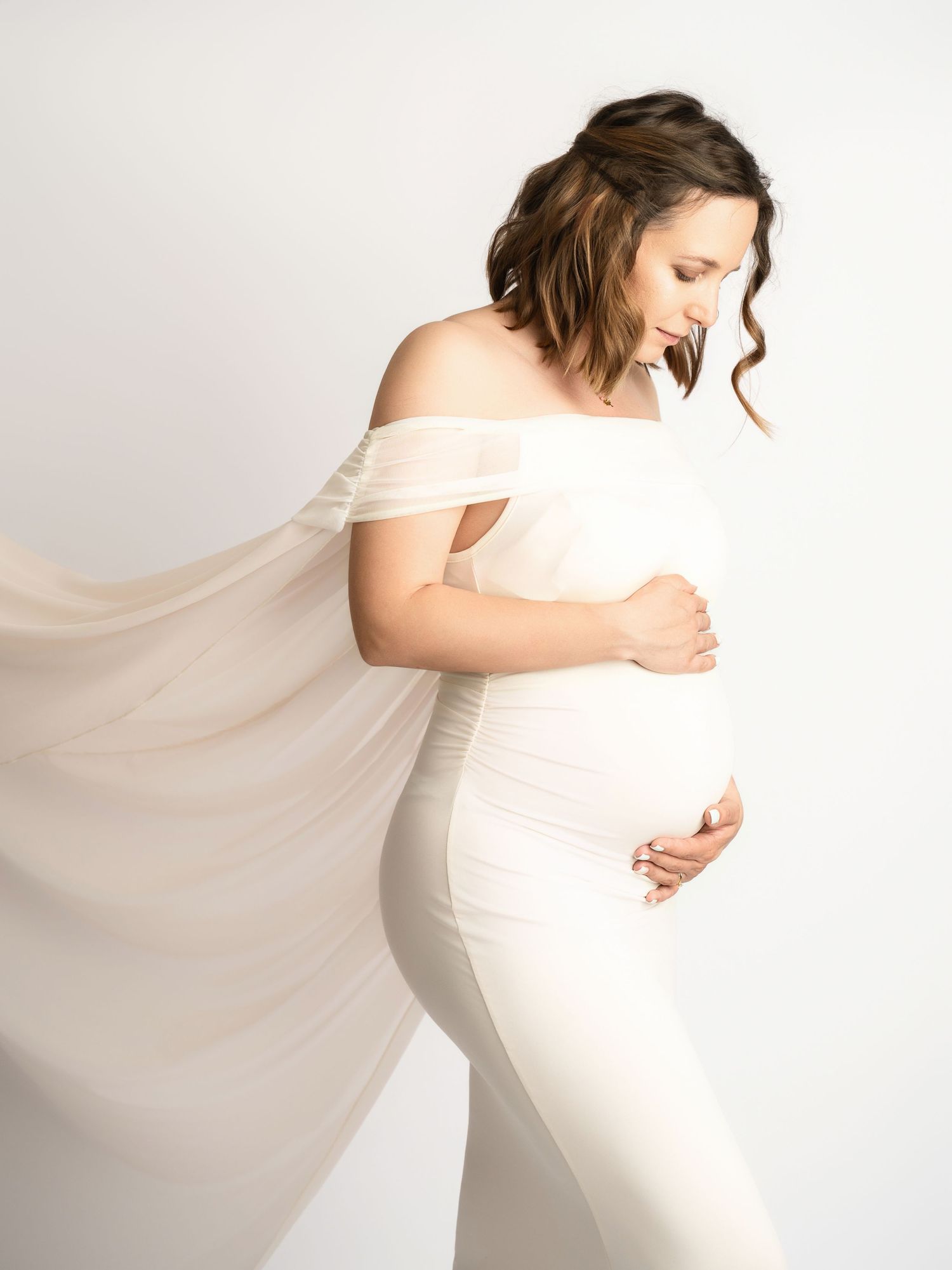 Pregnant woman in white off-shoulder fitted dress with sheer flowing fabric holding baby bump.