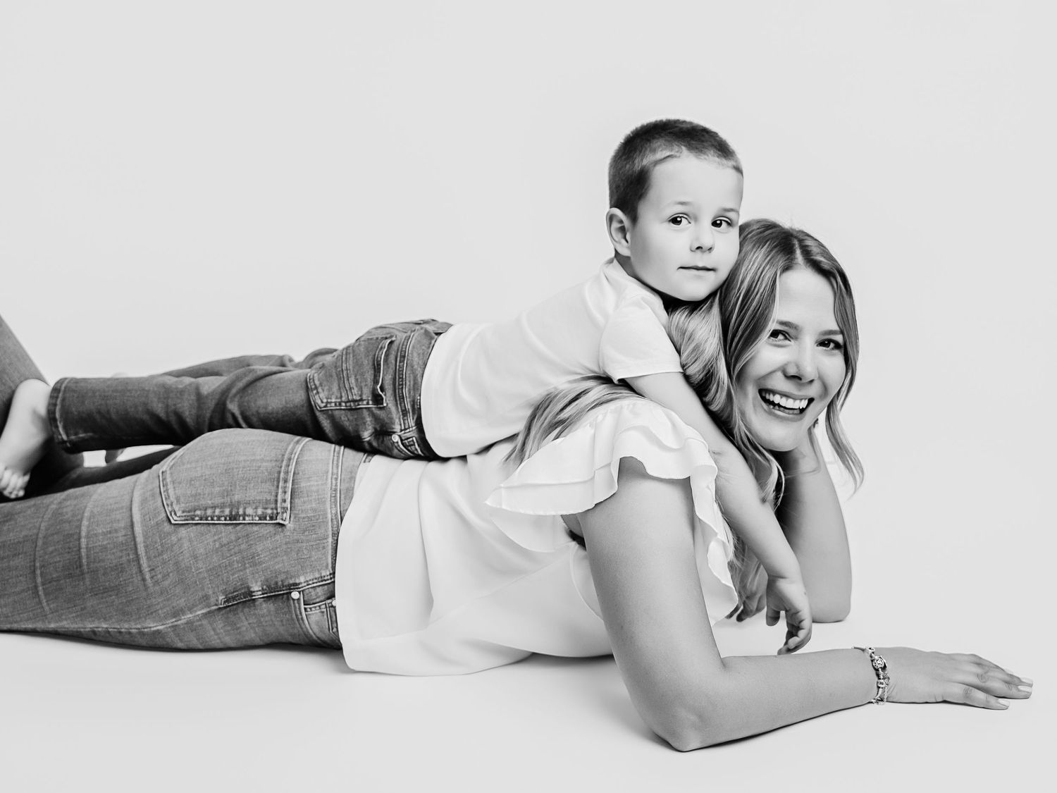 Black and white photo of a laughing mother lying down while her young son playfully rests on her back.