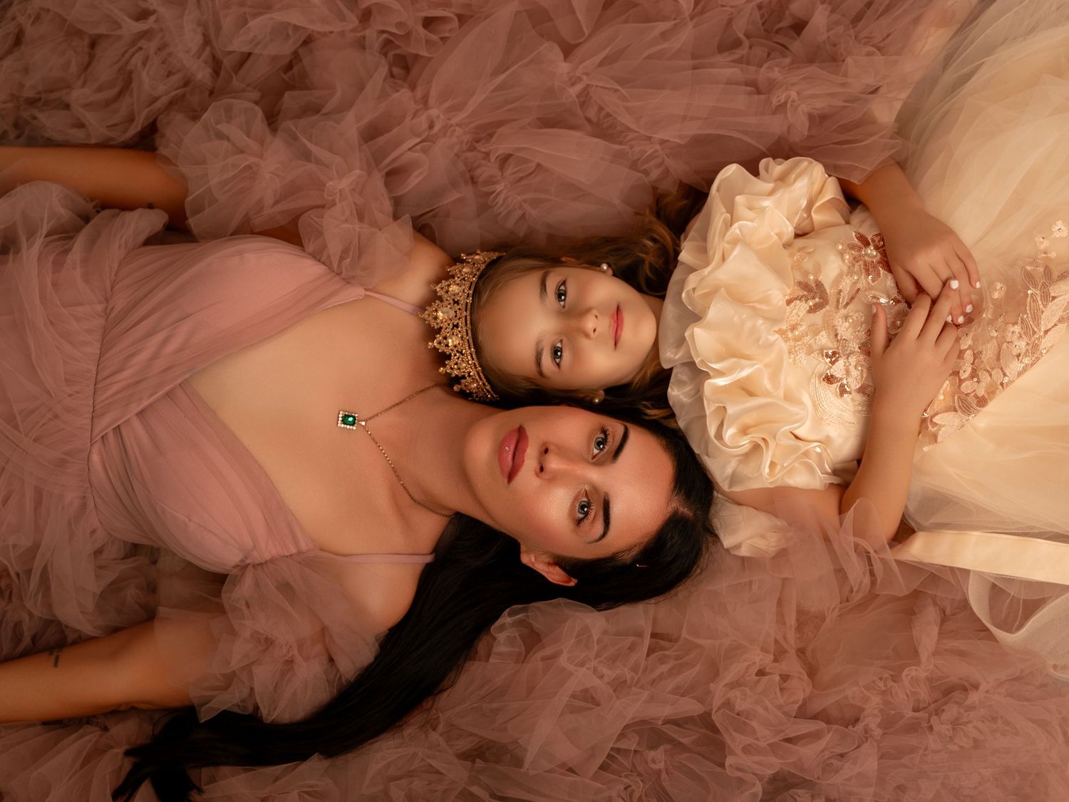 Two women in elegant pink tulle gowns lying together, wearing ornate jewelry in a dreamy, romantic portrait.