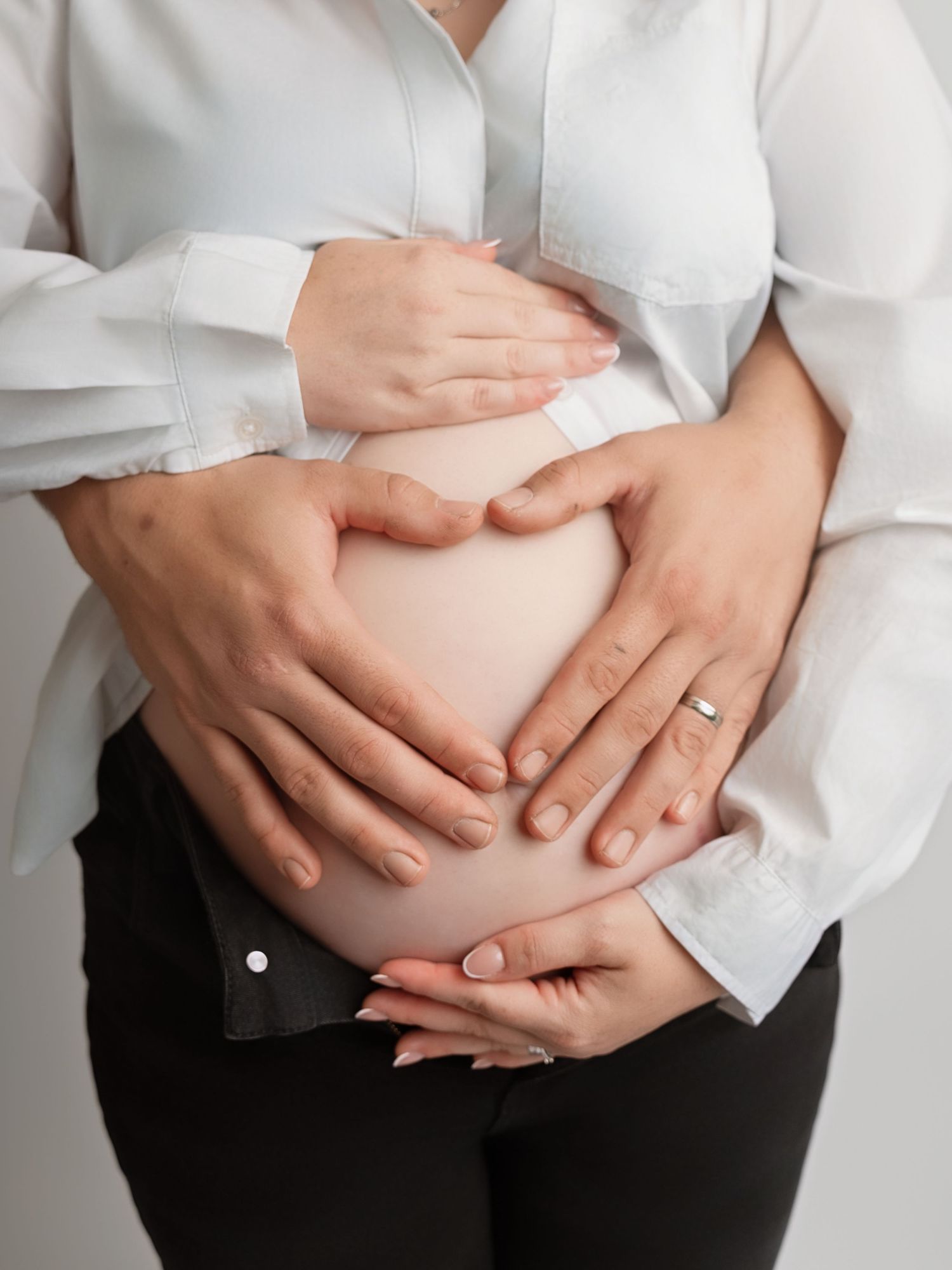 Pregnant woman in white shirt cradling baby bump with both hands forming heart shape, wearing engagement ring.