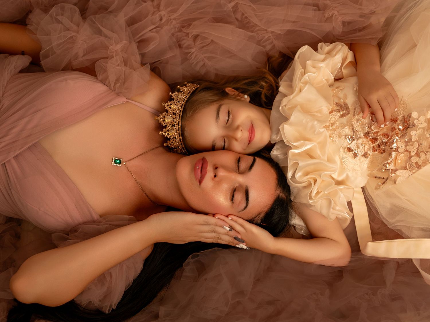 Two young women in elegant ball gowns and crowns lying together with eyes closed in a dreamy, romantic pose.