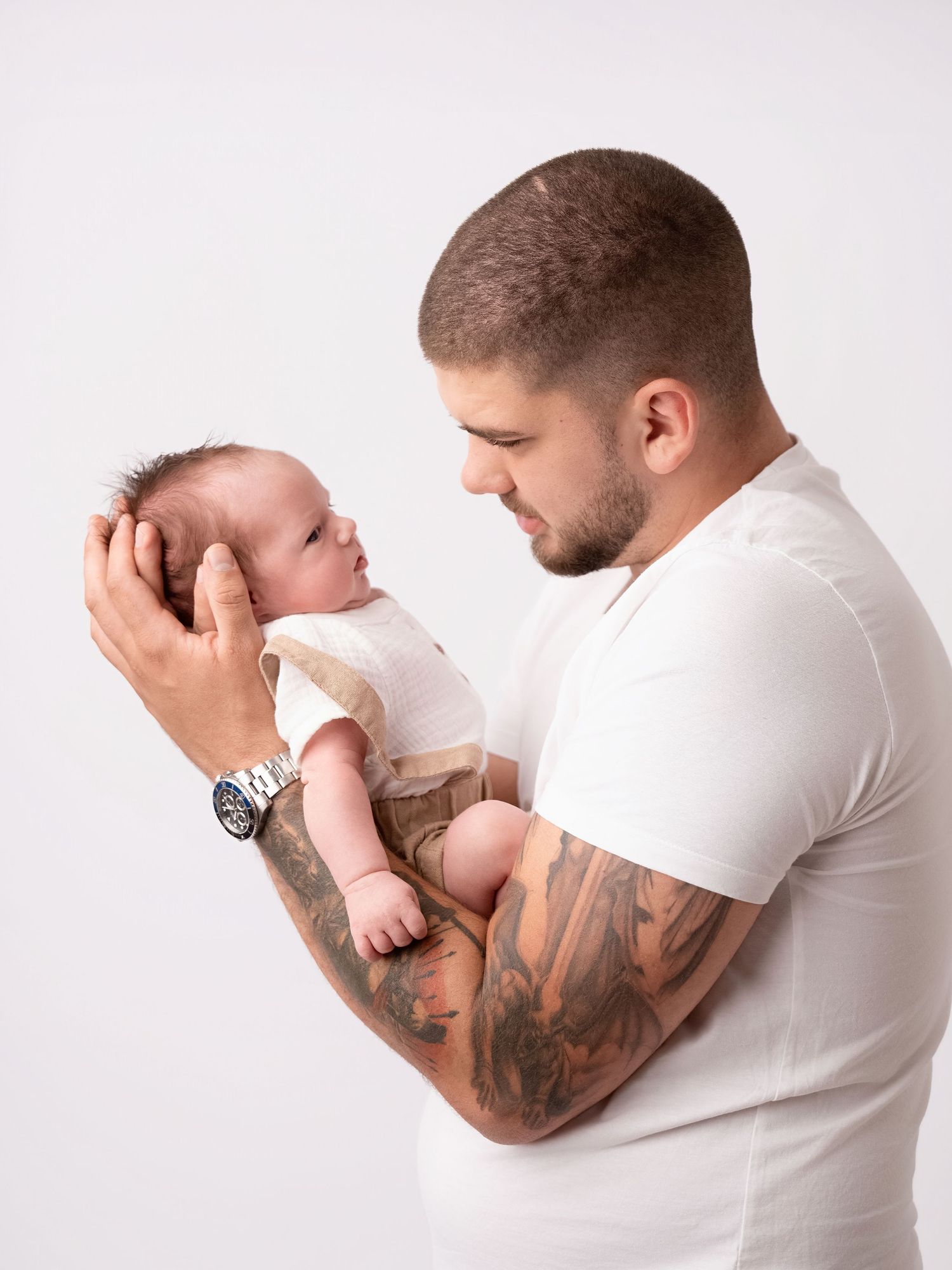 Tattooed father in white t-shirt gently holds newborn baby, gazing lovingly at infant against white background.