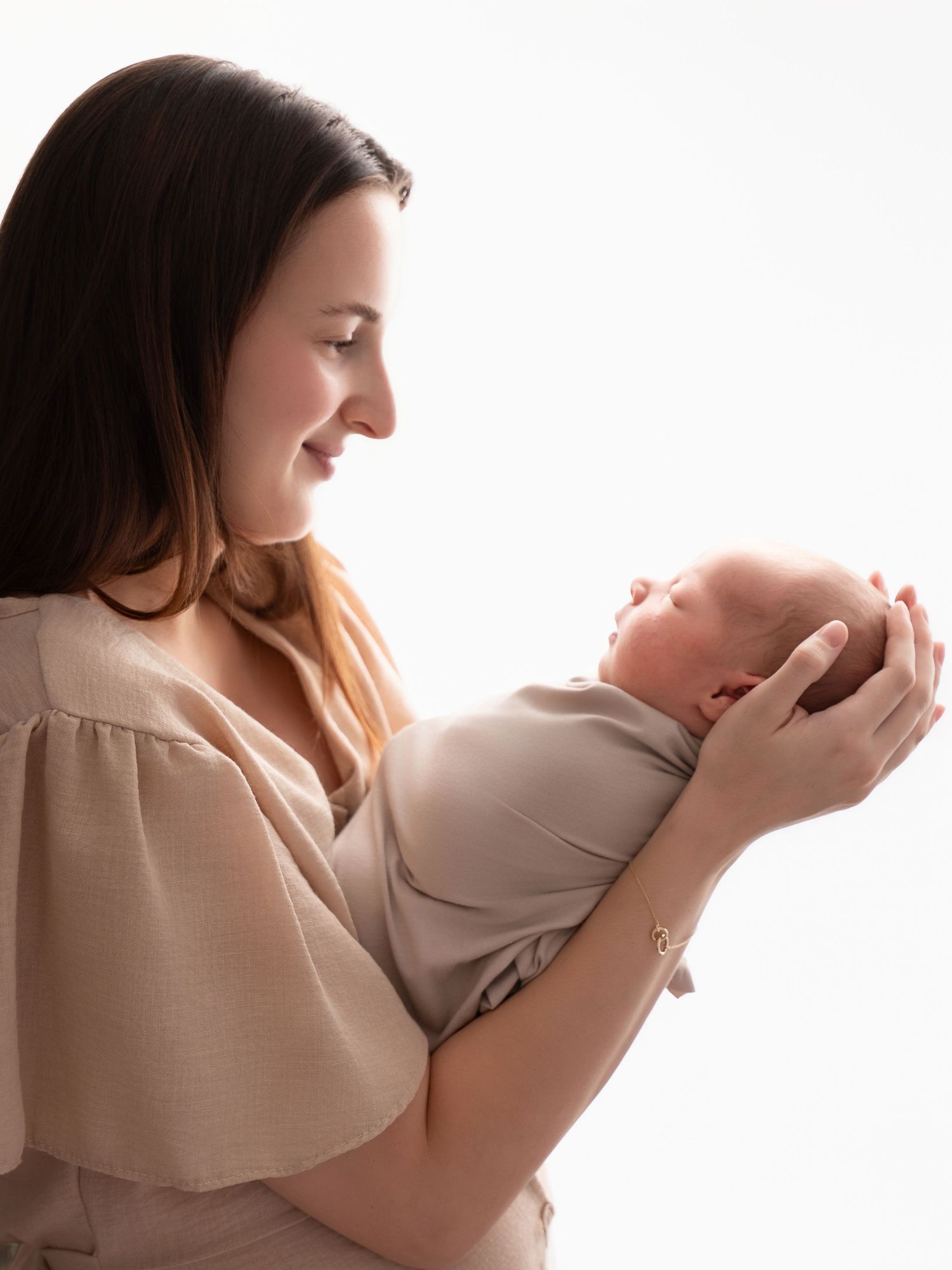 A mother tenderly holds her swaddled newborn baby in her hands, gazing lovingly at the infant against a white background.
