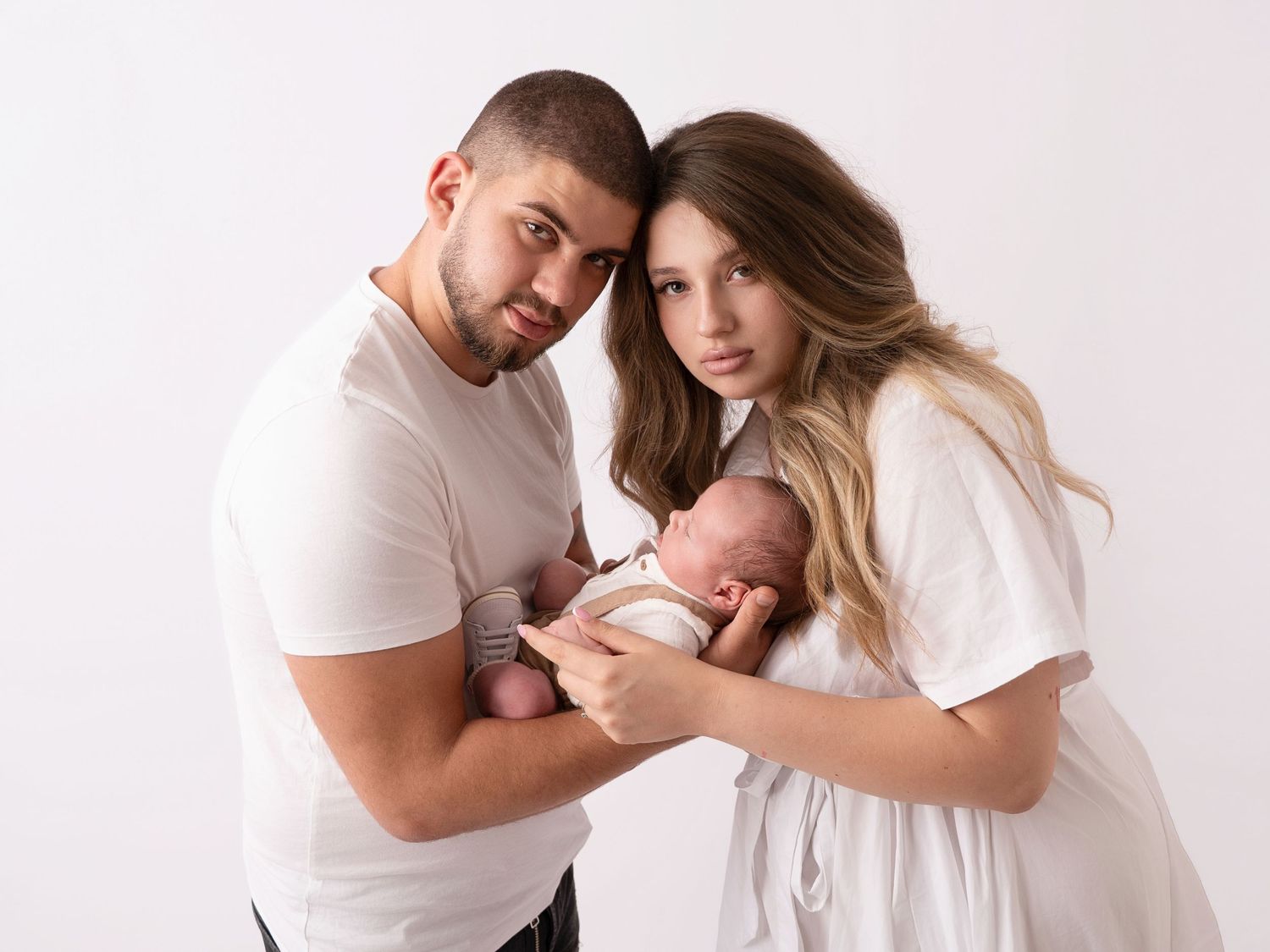 Young couple in white outfits tenderly holding their newborn baby against a soft white studio background.