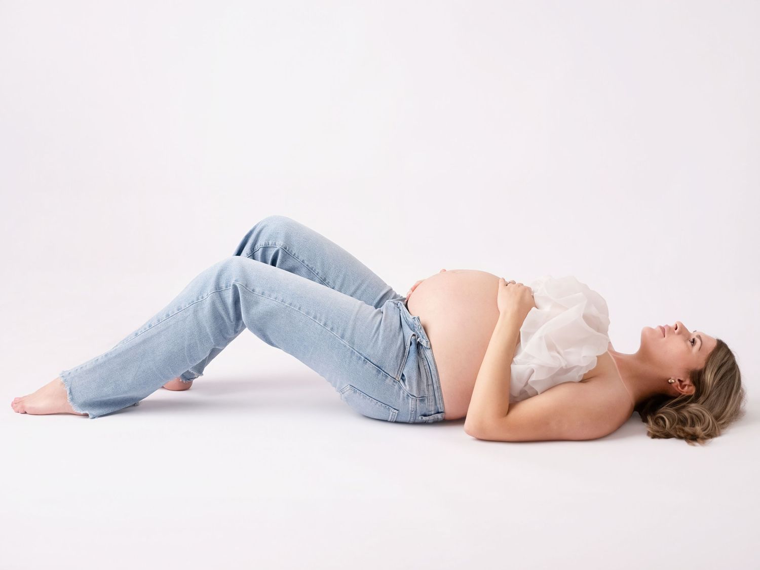 Pregnant woman lying on her back in light blue jeans and white top on a white background.