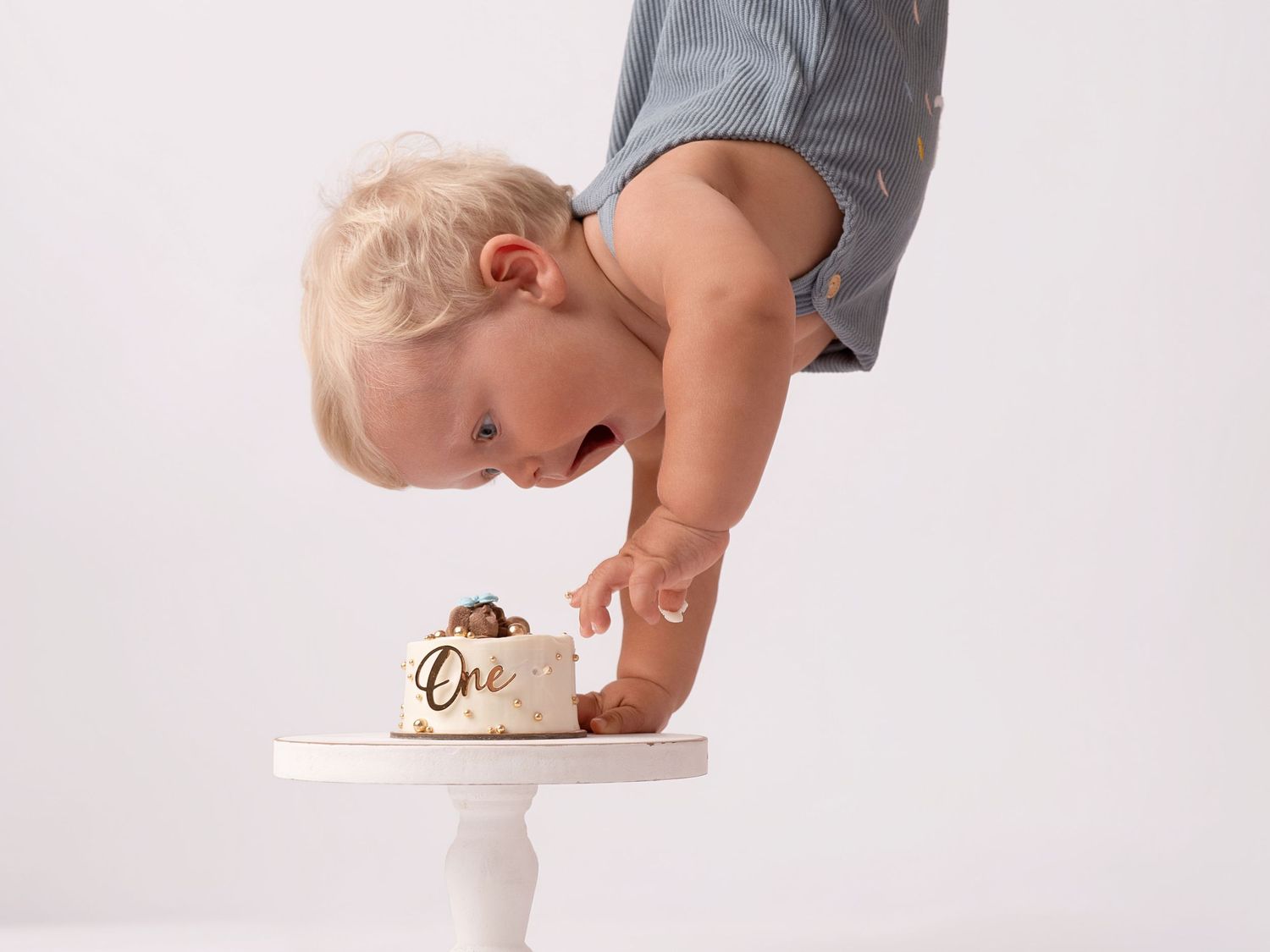 Blonde toddler in blue romper leaning over a small white cake stand with a first birthday smash cake labeled One.