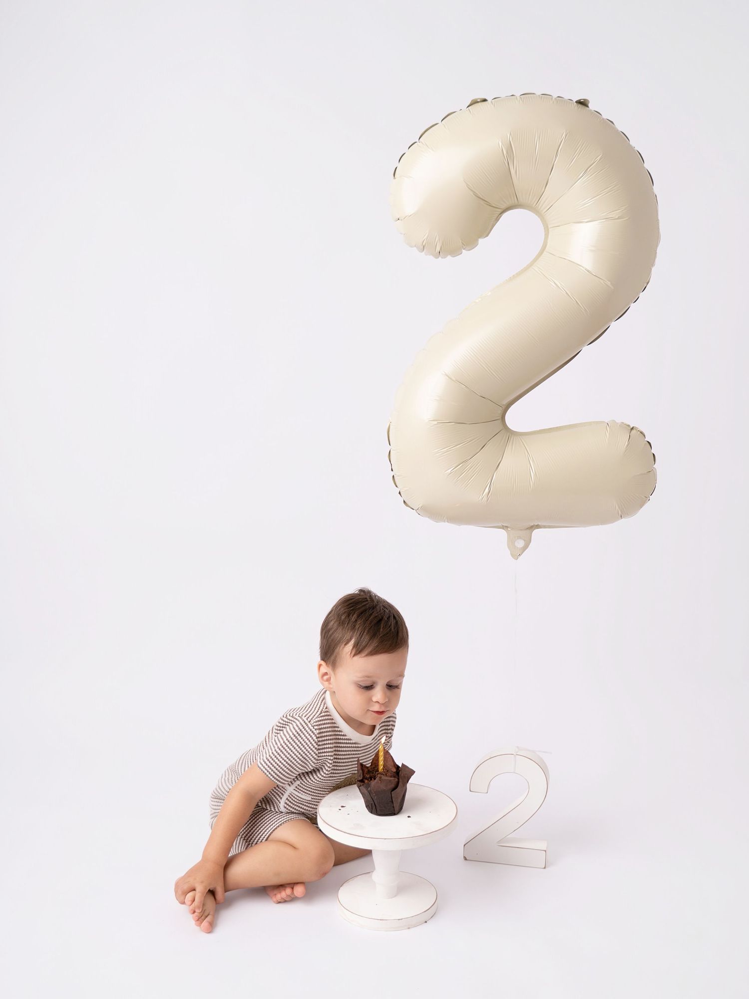 Toddler boy sitting on floor eating smash cake beside number 2 balloon and sign for second birthday photo.