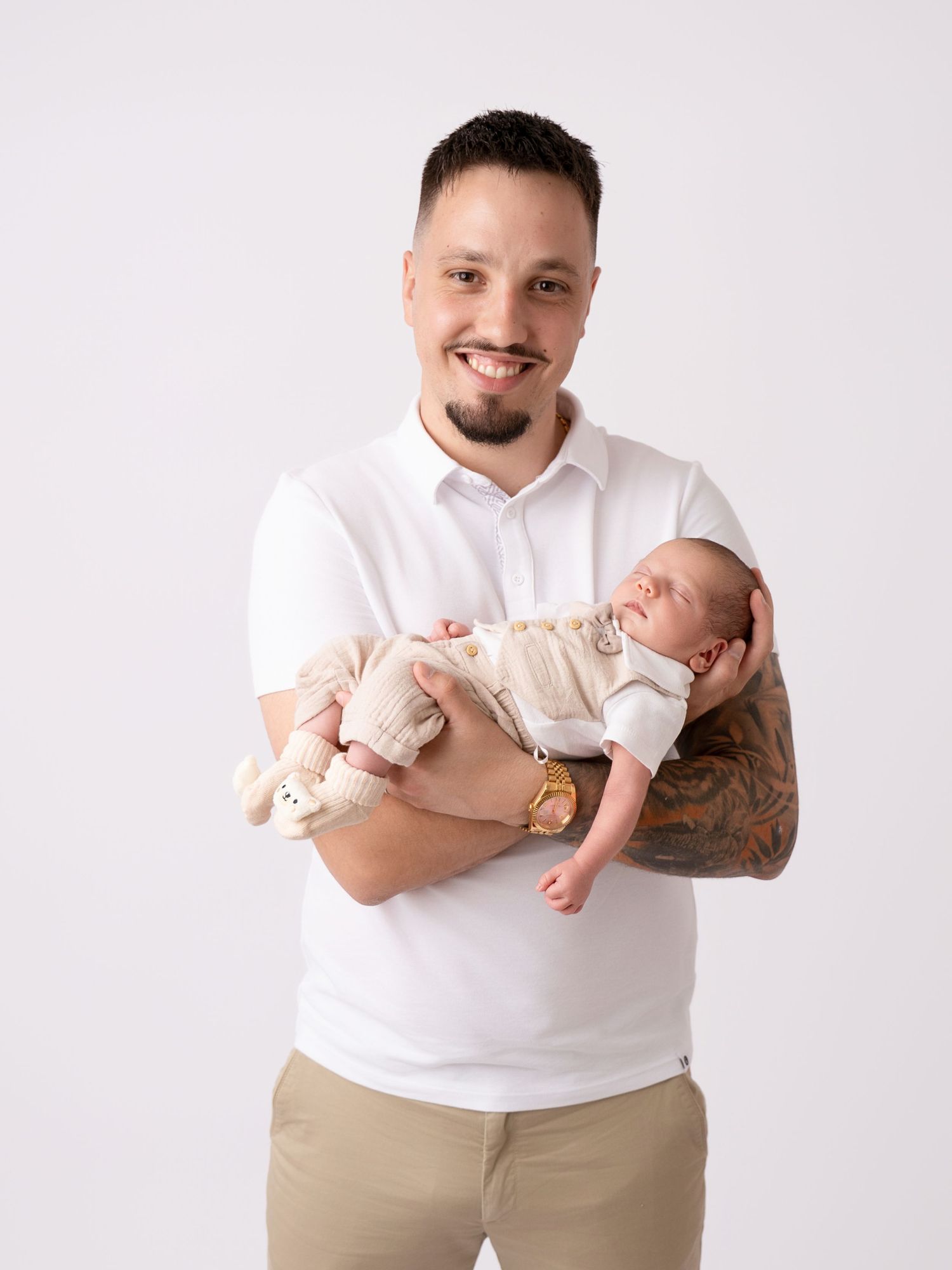 Smiling man in white polo shirt holding a sleeping newborn baby, with tattoos visible on his arm, against a white background.