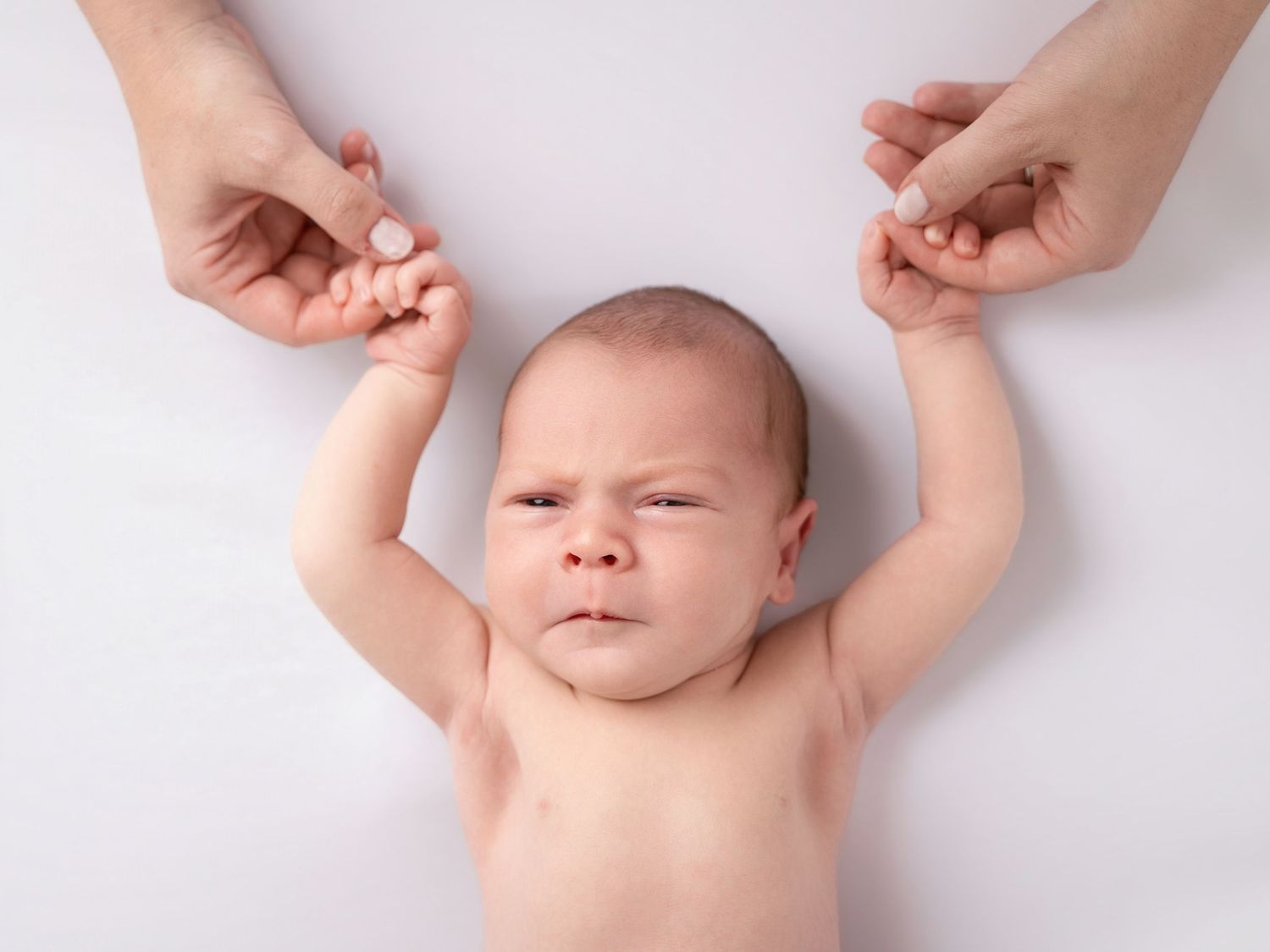 Newborn baby lying on white background holding adult fingers with both hands, looking up with a curious expression.