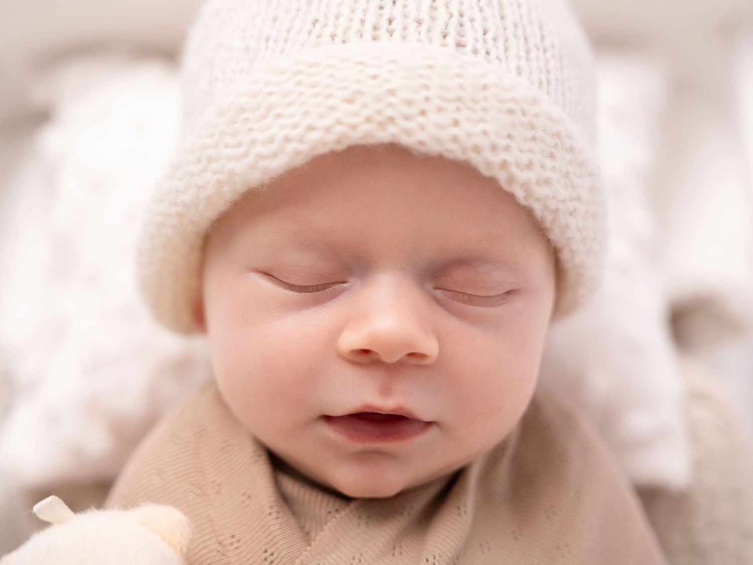 Peaceful newborn baby sleeping in a cream knit hat and soft wrap, close-up portrait photography.