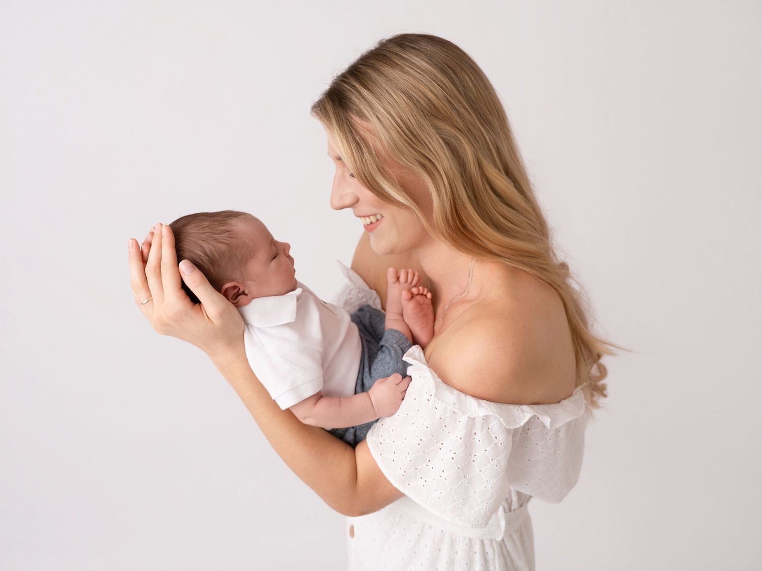A smiling mother in a white off-shoulder dress tenderly holds her newborn baby against a soft white background.