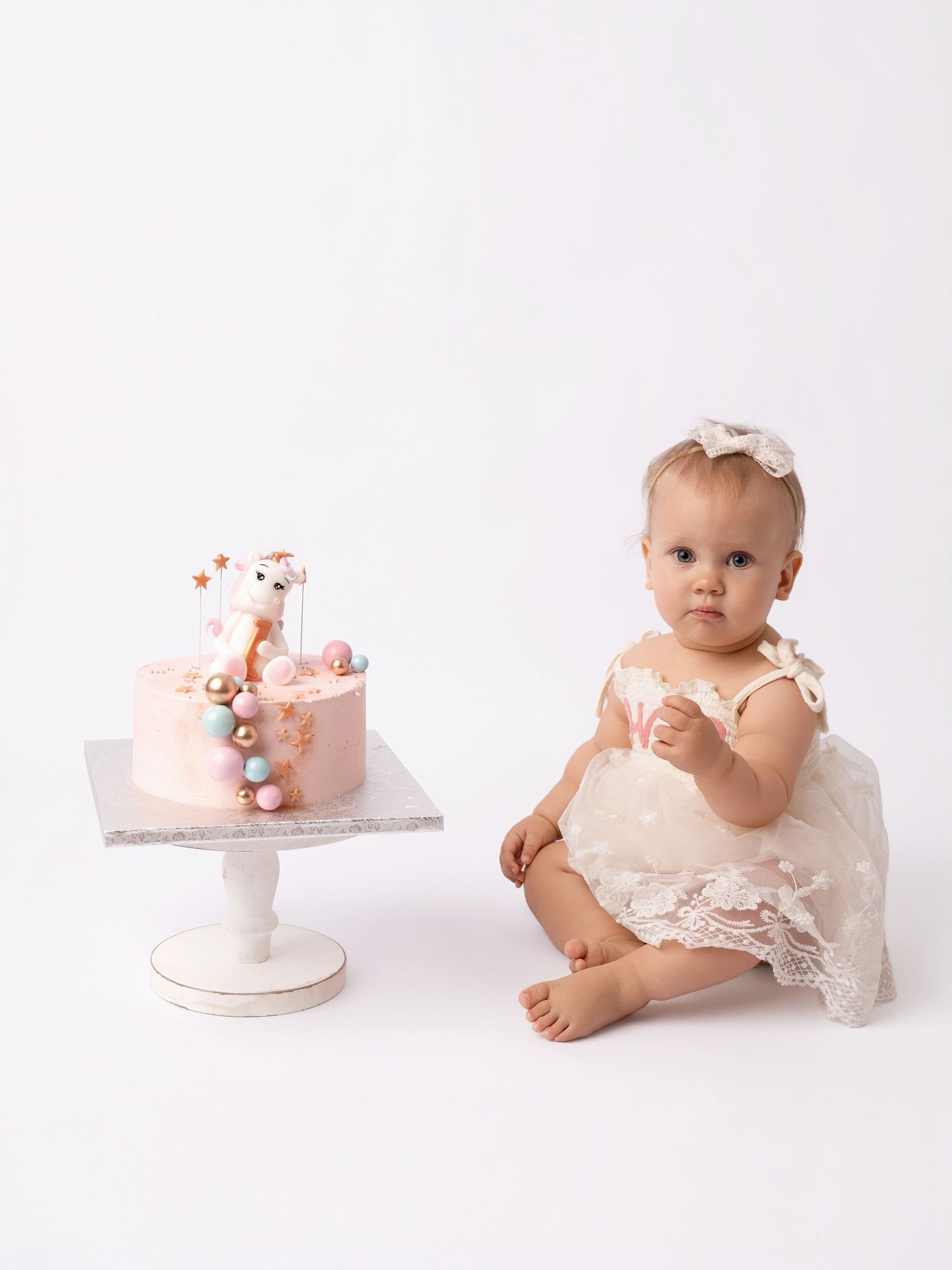 Baby girl in white lace dress sits beside a pink first birthday cake with giraffe topper on white background.