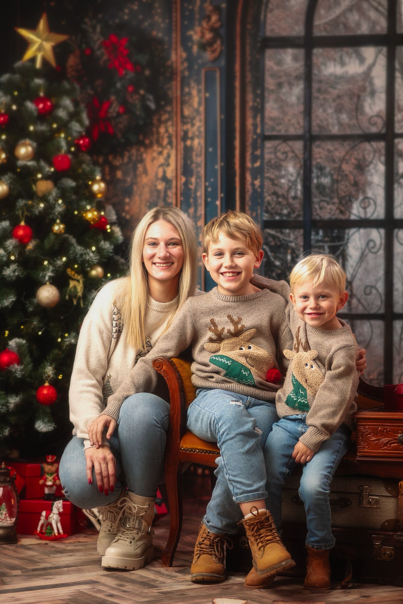 A smiling mother and two young boys in matching reindeer sweaters pose together by a decorated Christmas tree.