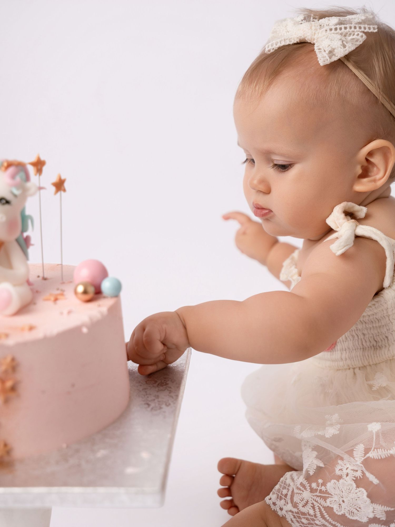 Adorable baby girl in white lace dress touches pink unicorn first birthday cake in studio setting.
