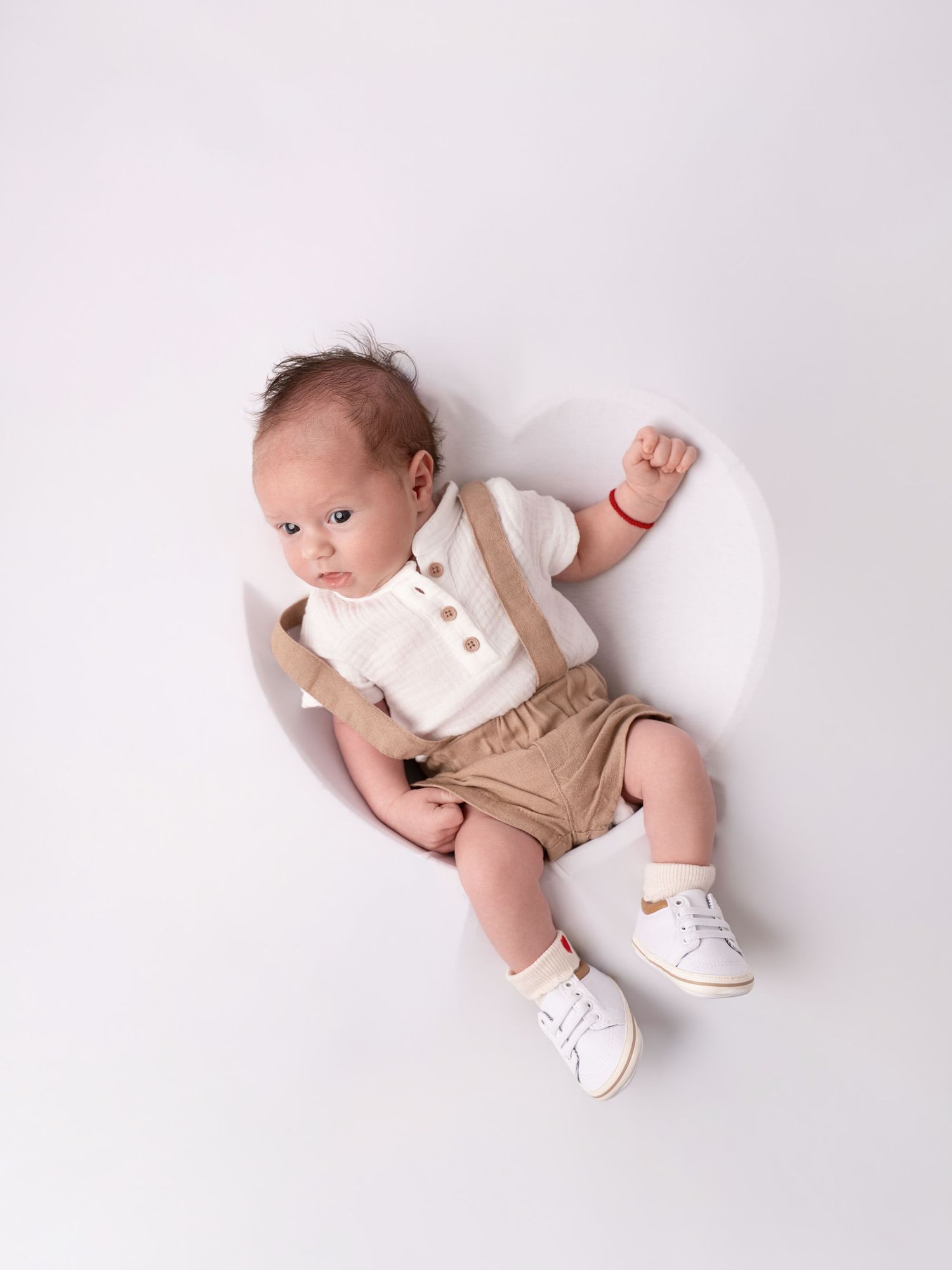 A cute baby boy in beige shorts with suspenders and white sneakers lying on a white background.