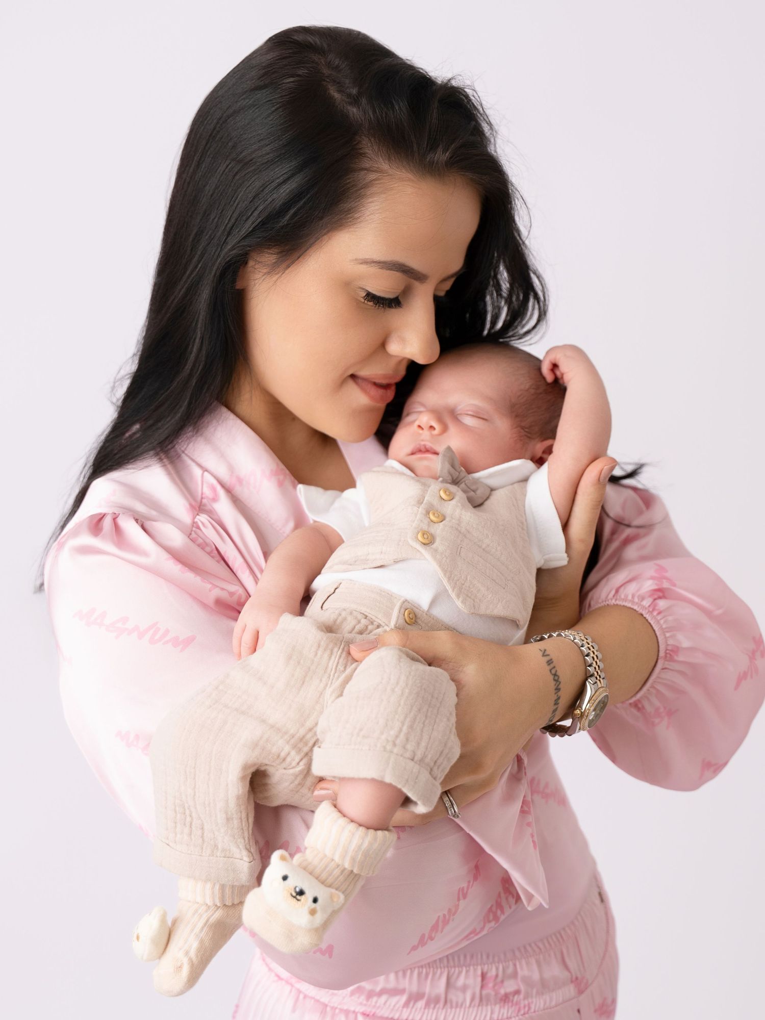 A young mother in pink holds her sleeping newborn baby close, touching foreheads tenderly in a studio portrait.