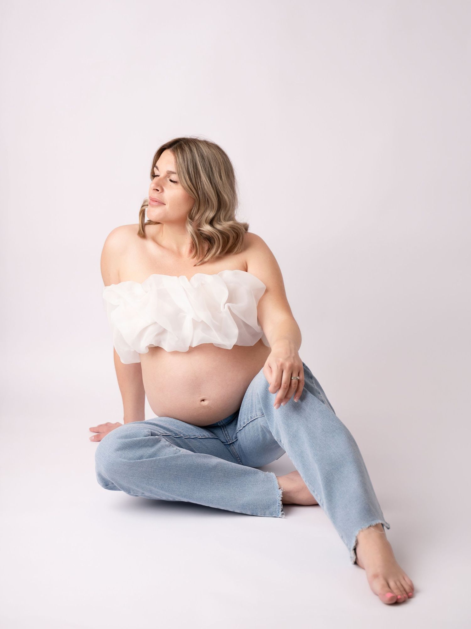 Pregnant woman in white ruffled tube top and light jeans sitting on floor, showcasing baby bump in studio maternity photo.