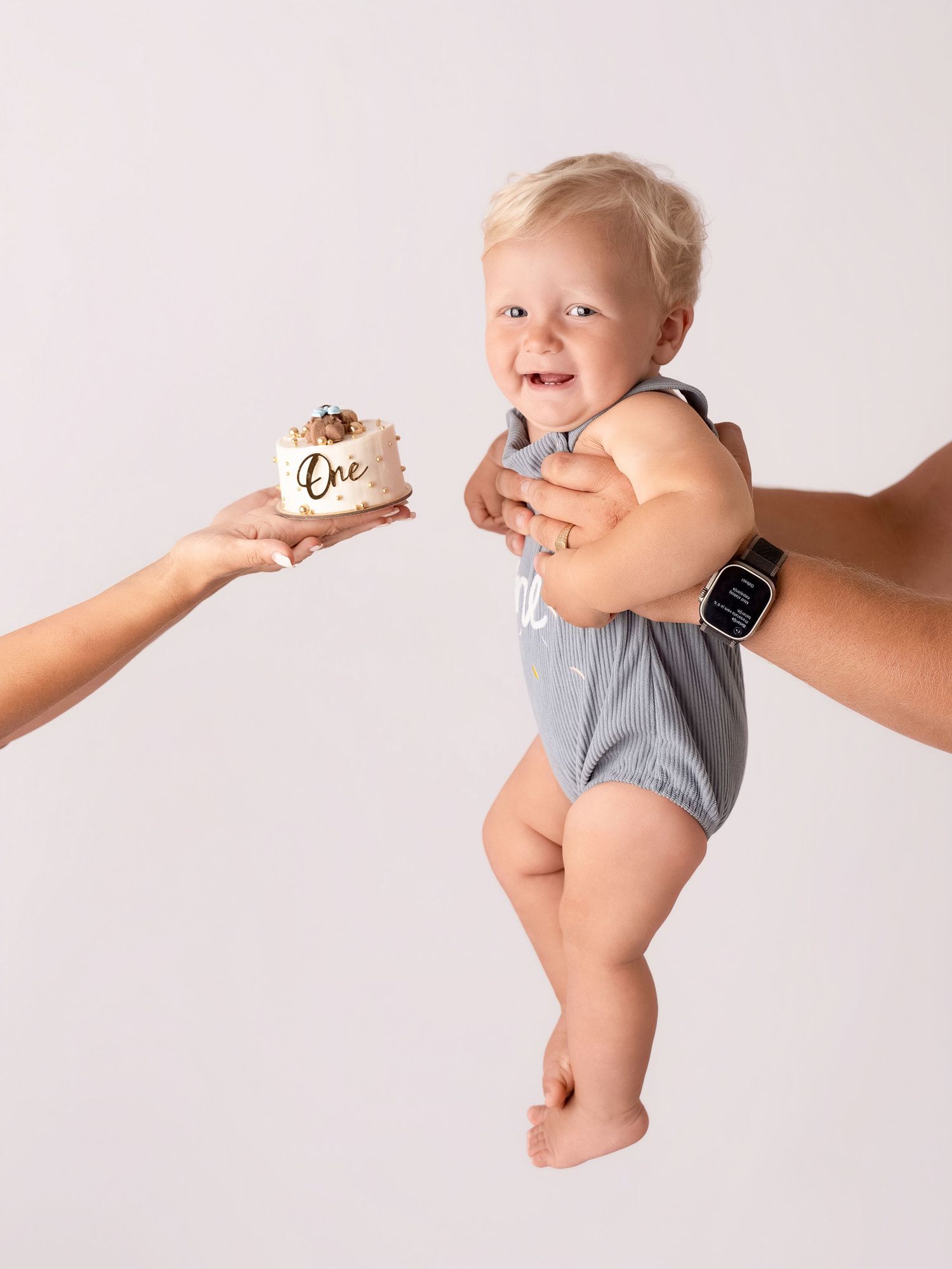 A smiling baby in a gray romper reaches for a first birthday smash cake labeled One while being held up by an adult.