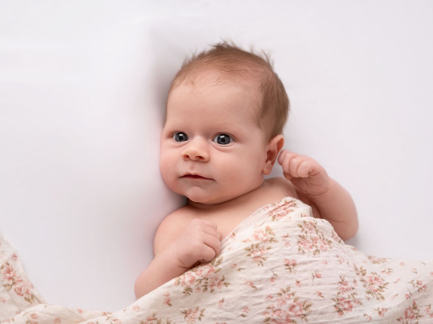 Newborn baby wrapped in a floral muslin swaddle blanket lying on a white background.
