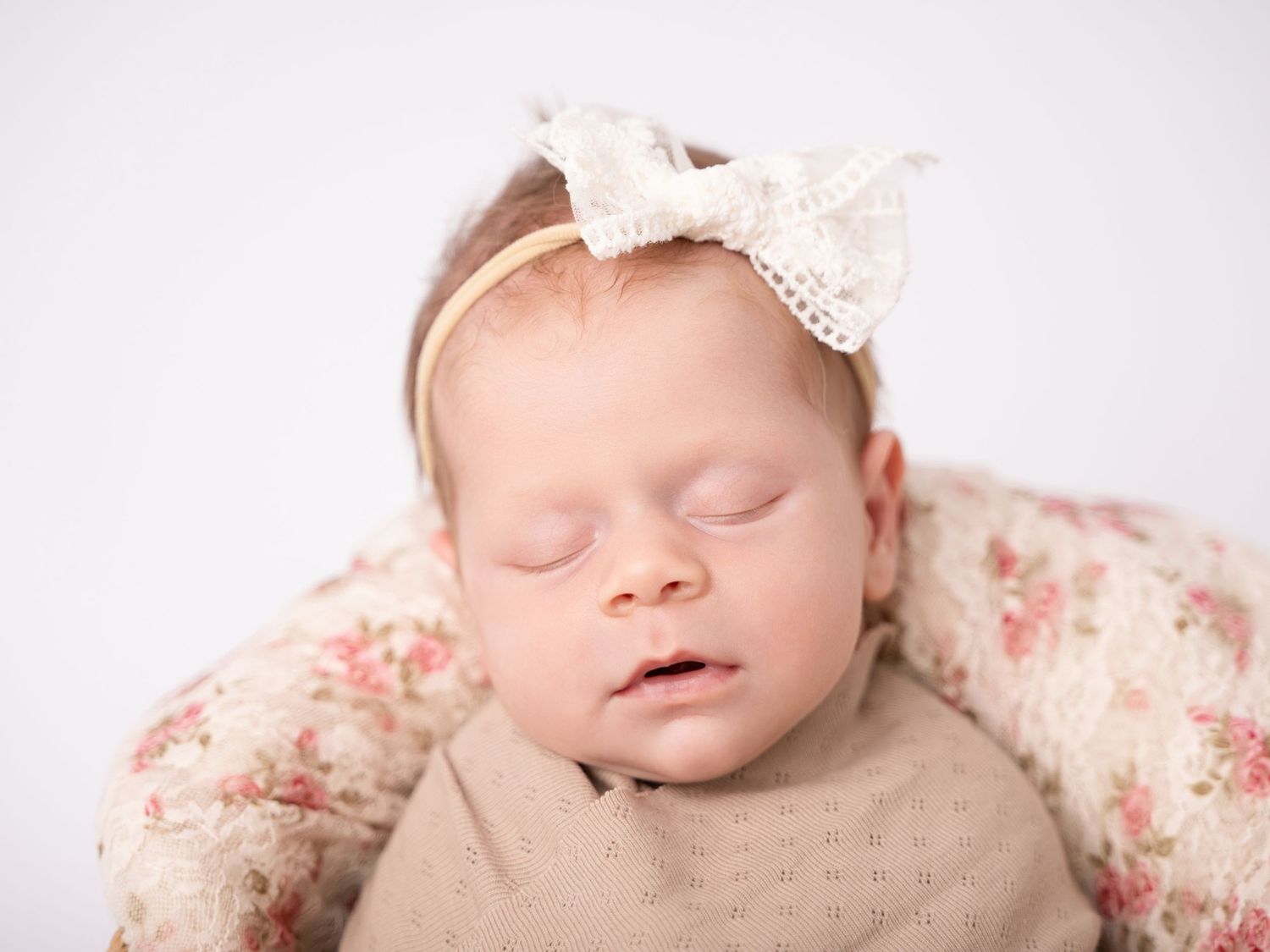 Sleeping newborn baby girl wearing a lace bow headband, wrapped in a floral blanket during a professional newborn photo session.