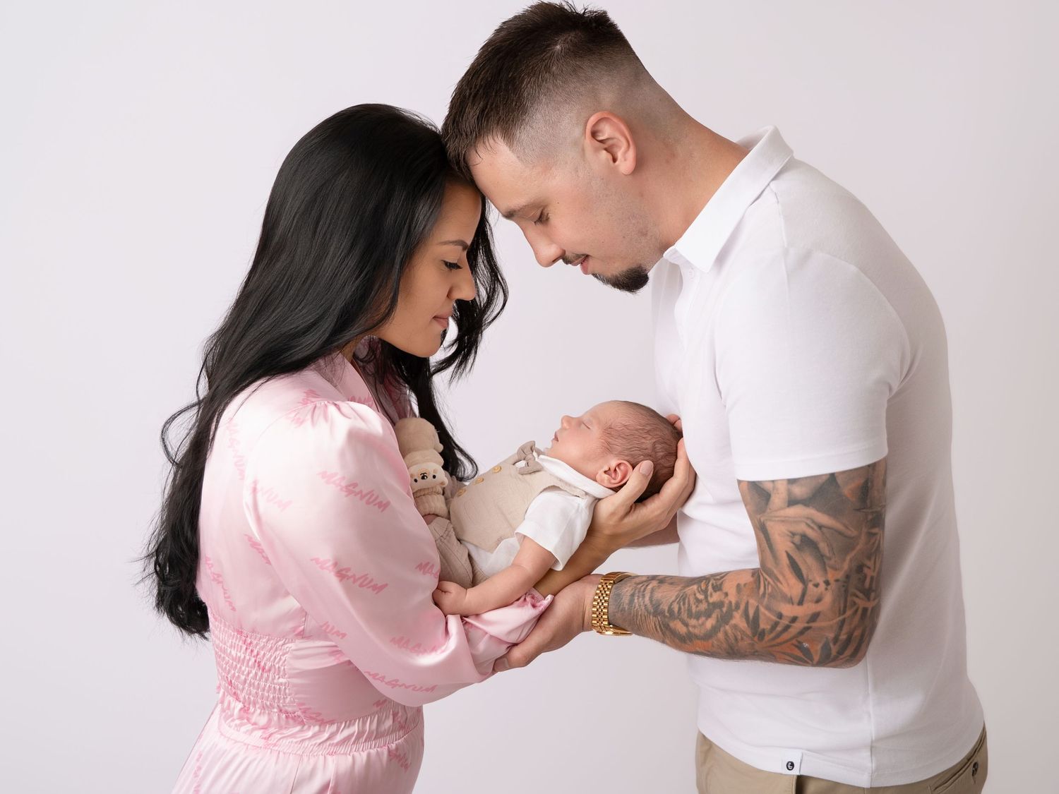 A couple in a photography studio tenderly cradles their newborn baby, the mother in pink and father in white with tattoos.