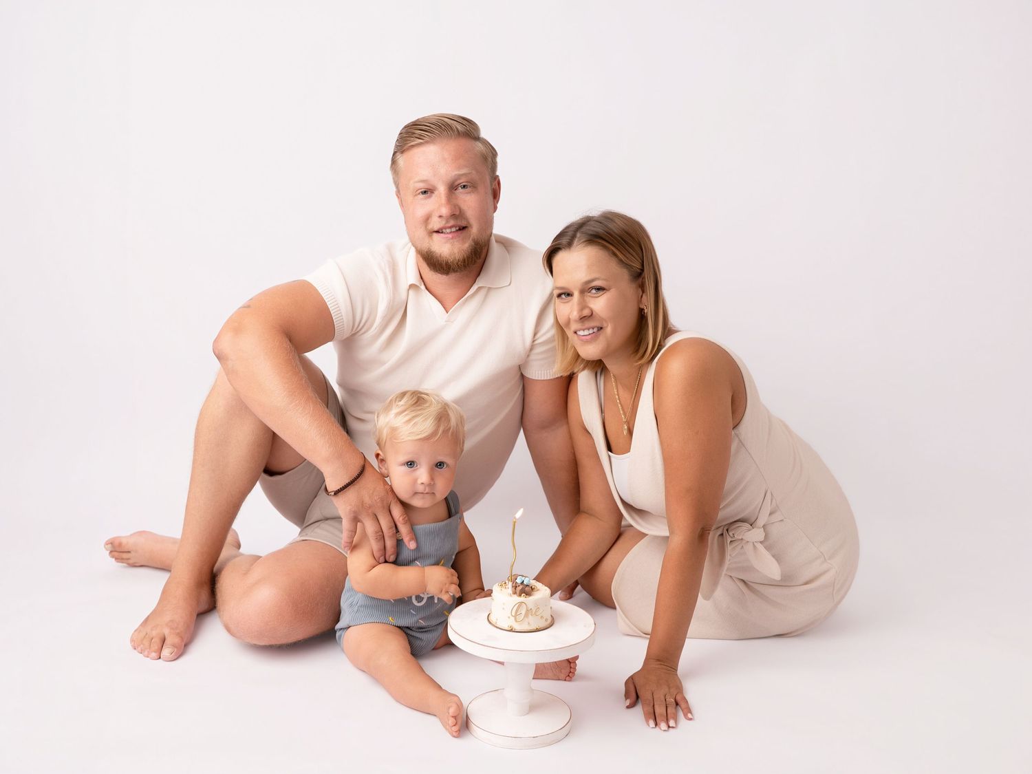 Family of three posing with a small birthday cake on white background, parents smiling beside their toddler.