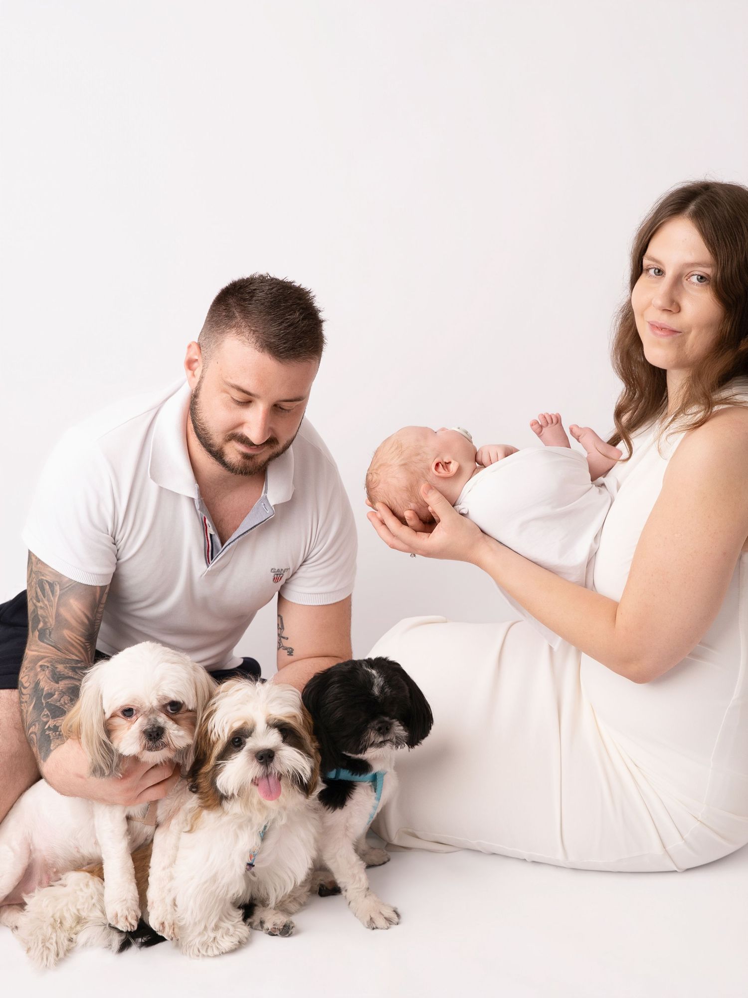 New parents holding newborn baby with three small dogs in a bright white studio family portrait photo.