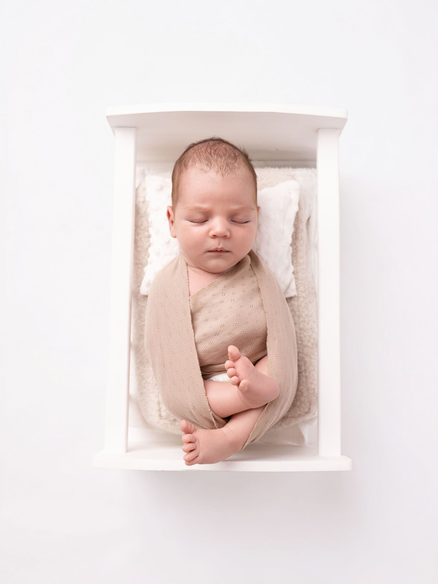 Newborn baby sleeping peacefully in a small white wooden crib, wrapped in a soft beige swaddle on a white background.