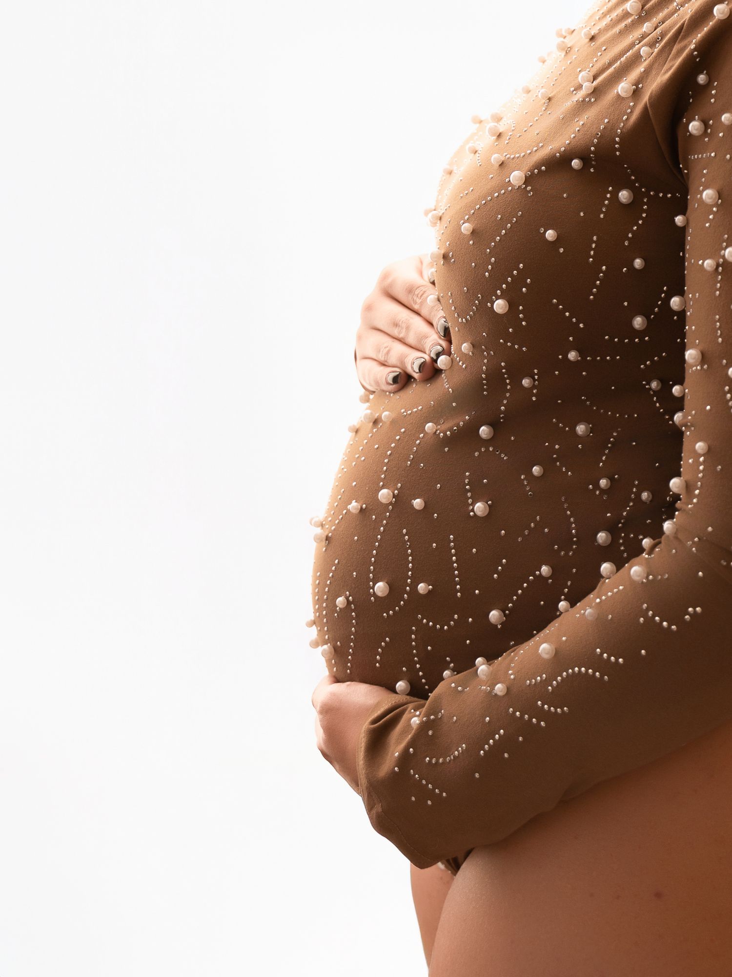 Pregnant woman in a brown pearl-embellished sheer bodysuit cradling her baby bump against a white background.